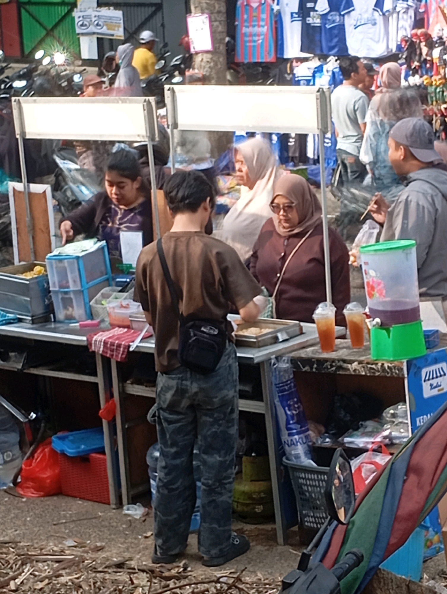 In the city of Cimahi, Indonesia, a street food vendor operates from a mobile cart. The cart is laden with various food items and drinks, including what appears to be a large dispenser of purple liquid, likely a beverage. Two individuals stand near the cart, engaged in a transaction or conversation, while others mill about in the background. The scene is characteristic of a bustling street market, with numerous stalls and people present. The overall atmosphere is lively and dynamic, reflecting the vibrant street culture of Cimahi.
