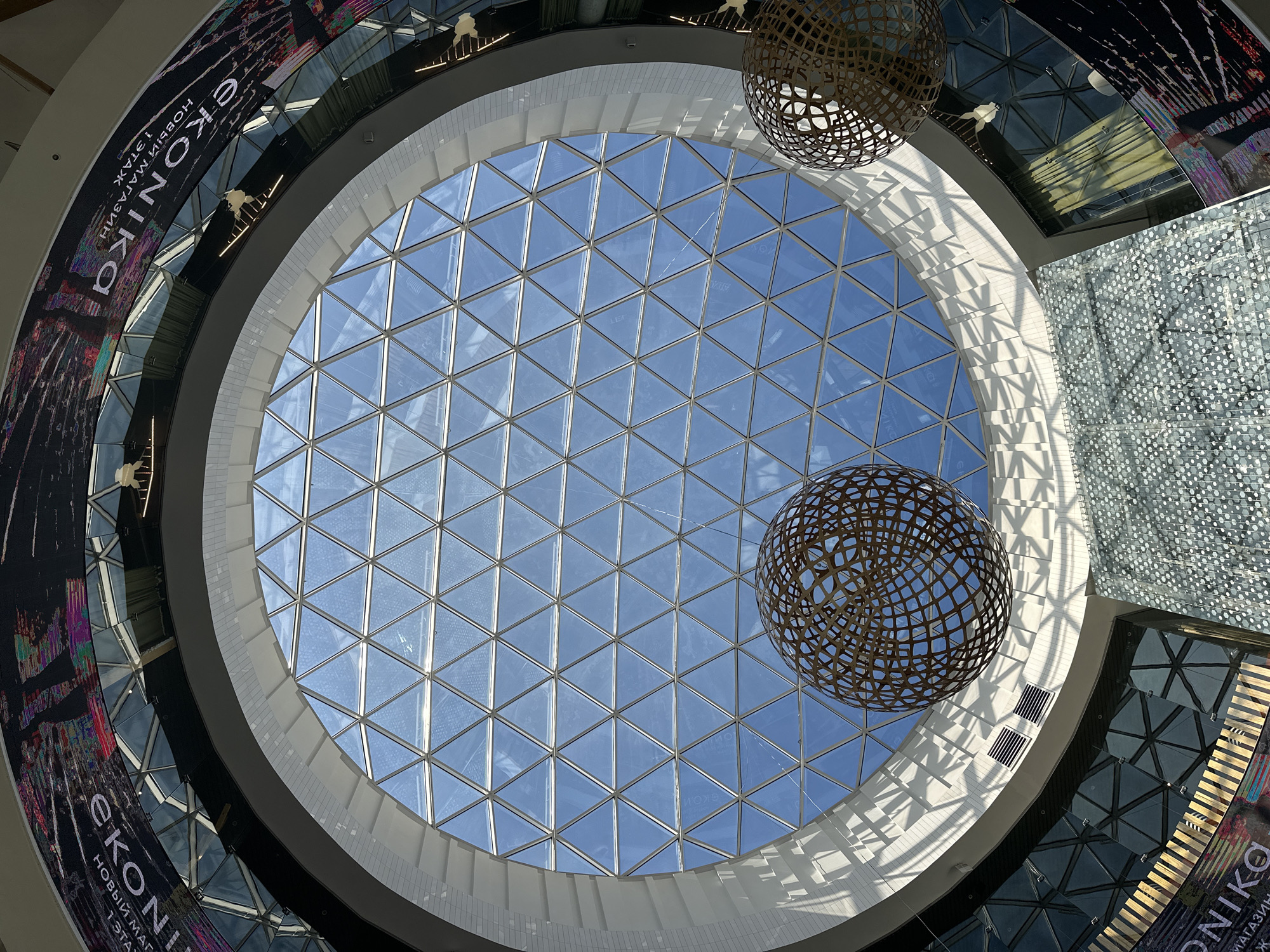 This image captures a low-angle perspective looking up at the grand interior of a modern building, likely a shopping mall in Kazan, Russia. The central focus is a large, circular geodesic skylight made of numerous triangular glass panels, revealing a clear blue sky and bright daylight.
Two prominent, spherical, open-work decorative fixtures or sculptures hang from the ceiling, one in the upper right and another in the lower right quadrant of the image. These lattice-patterned spheres cast intricate shadows onto the curved white architectural surfaces below, which feature vertical grooves and steps.
Surrounding the skylight is a multi-level circular structure with various materials and textures. A large, curved digital display or billboard is visible on the left and wraps around the structure. Prominently displayed in Cyrillic is the word "ЭКОНИКА" (EKONIKA), followed by "НОВЫЙ МАГАЗИН" (NEW STORE) and "1 ЭТАЖ" (1ST FLOOR), indicating a retail space. Other visible architectural elements include reflective panels and a textured wall with a grid-like or pixelated pattern on the right side. The overall scene is devoid of people or specific activities, emphasizing the architectural design and the interplay of natural light and shadows within the spacious interior.