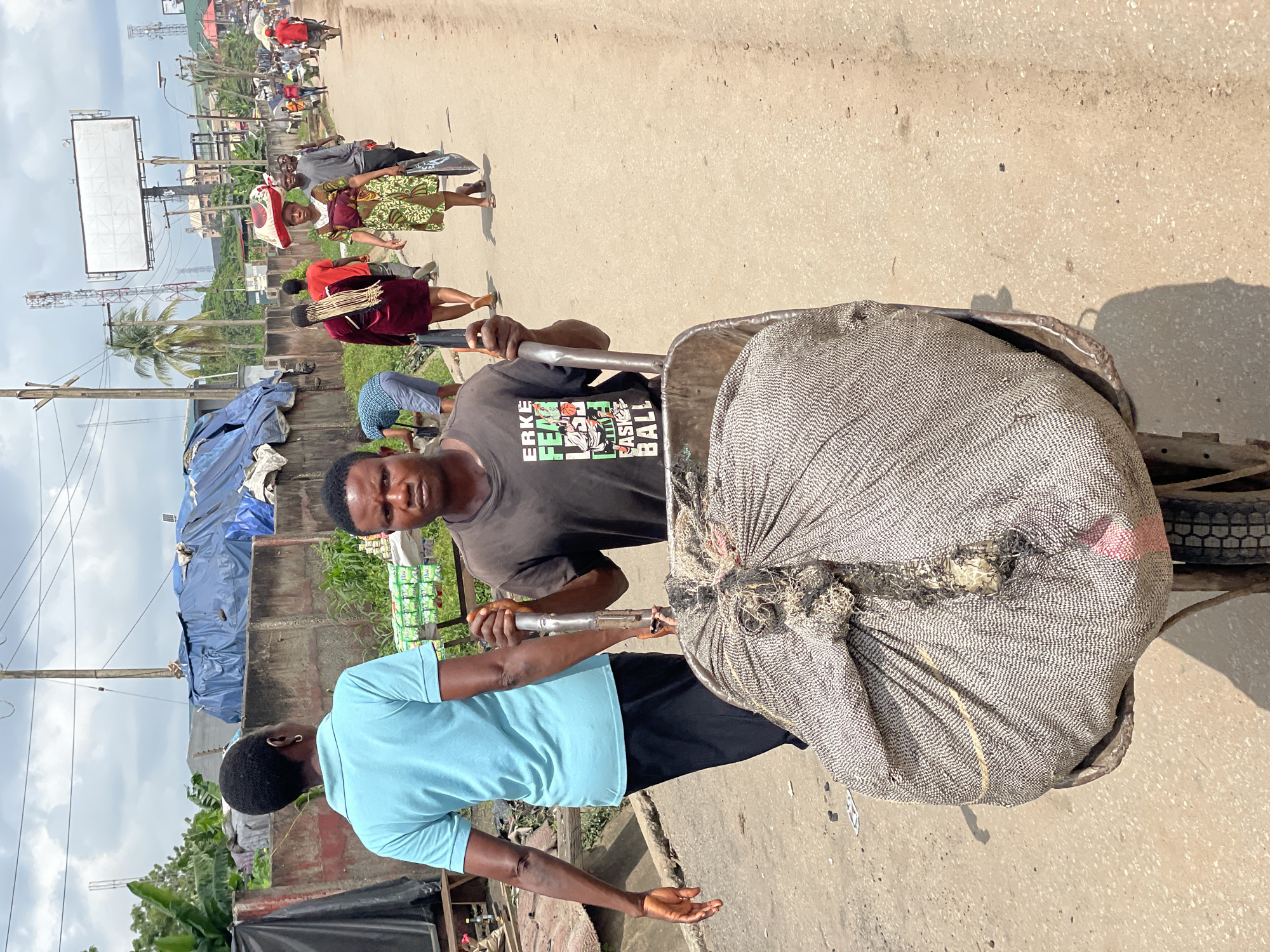 A man in a black t-shirt with graphics on it is pushing a wheelbarrow filled with a large, rough-textured sack on a dusty road in Fegge, Nigeria. The man is looking directly at the camera. A person in a light blue t-shirt and black pants walks in front of him and slightly to his left, with their back to the camera. In the background, other people are walking along the road. Buildings, tarpaulin-covered structures, and utility poles are visible. The sky is cloudy.