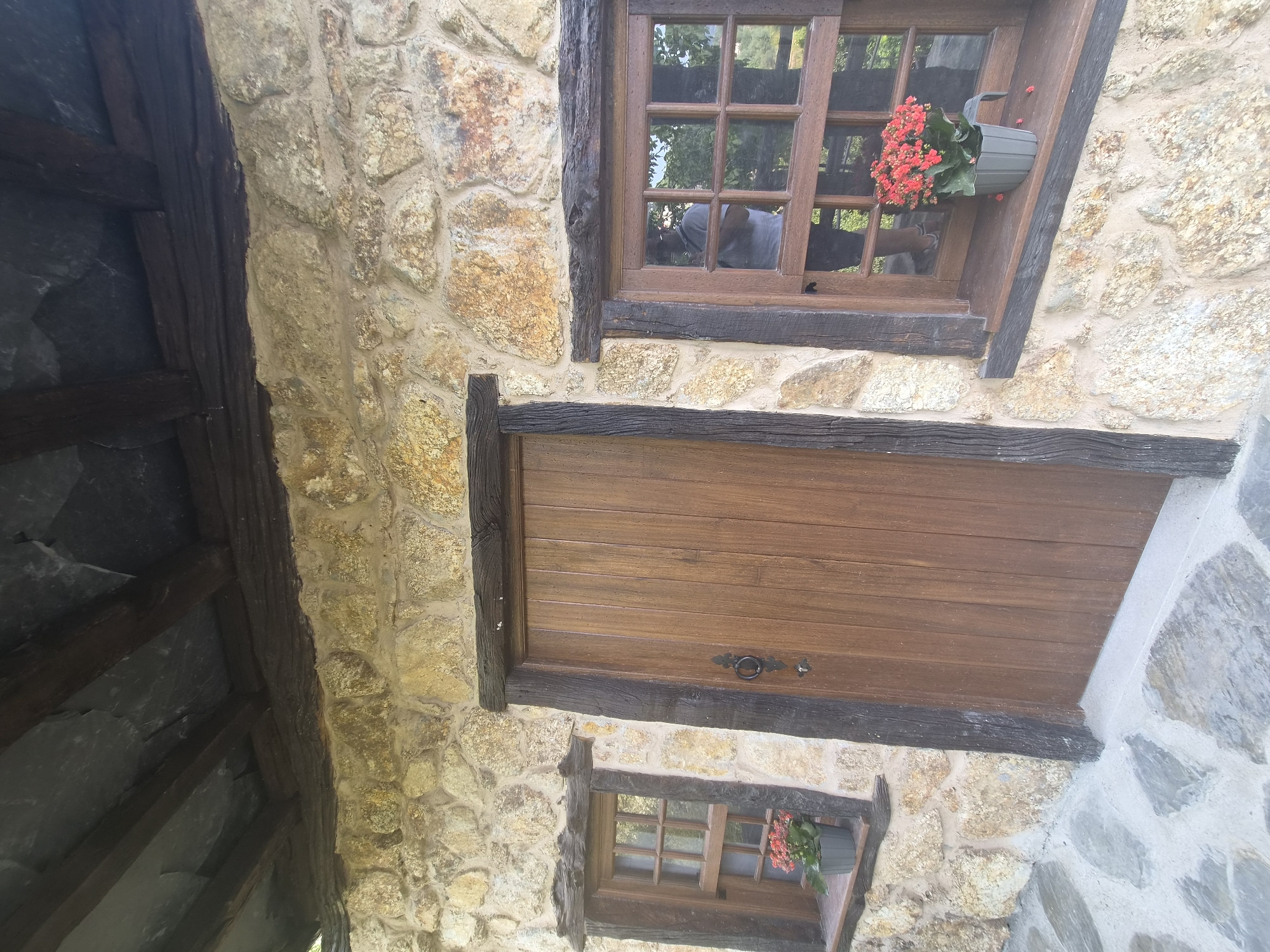 The image displays the rustic exterior of a stone building, likely a house or cottage, in Seia, Portugal. The stone walls are constructed from irregular, light-colored rocks with visible mortar. A weathered wooden door, vertically paneled, is centrally positioned, featuring a decorative black metal door handle. To the left of the door is a small, wooden-framed window with multiple panes, housing a potted plant with red flowers on its windowsill. To the right of the door, a larger, similar wooden-framed window reveals a reflection of a person wearing a light-colored shirt. This window also has a windowsill adorned with a potted plant bearing red flowers. Above the stone wall, dark wooden beams support a roof covered with dark slate tiles. The ground in front of the building is paved with irregular, grey flagstones. The scene evokes a sense of traditional architecture and rural charm.