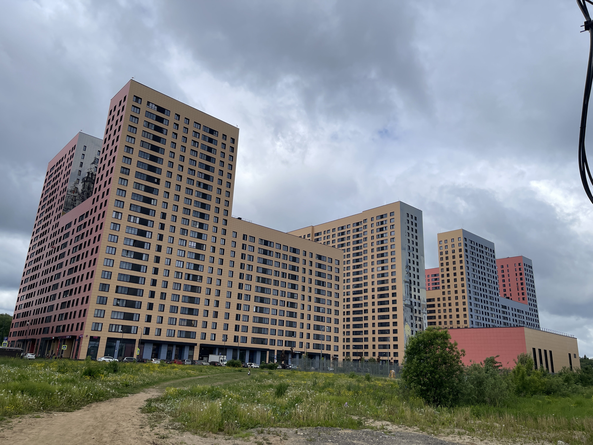The image shows a cluster of recently constructed residential buildings in Kommunarka, Russia. The buildings are multi-story and characterized by a modern, block-like architectural style with predominantly square windows. Facade colors vary across the buildings, including shades of beige, pink, gray, and blue. Several buildings are interconnected, forming a cohesive architectural unit.
In the foreground, there is a dirt path leading towards the buildings, bordered by a grassy field with scattered wildflowers. Some cars are visible parked near the base of the buildings. A chain-link fence separates the field from the building complex. Trees and shrubbery are also present in the foreground, adding to the green space around the buildings. The sky is overcast with gray clouds, suggesting a cloudy day. A few utility wires are visible in the upper right corner. There are no people visible in the image.