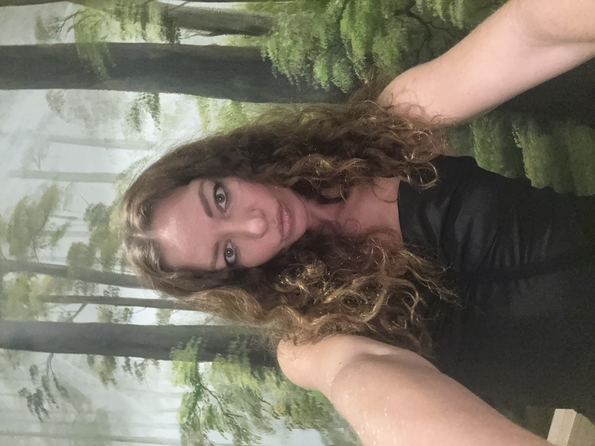 This is a close-up, vertically oriented selfie of a woman with long, curly brown hair. She is wearing a black top and is looking towards the camera with a slight smile. Her hair is loose and frames her face, with some strands falling onto her shoulders. The background appears to be a mural or backdrop depicting a forest scene with trees and green foliage, with a soft, hazy effect. The lighting suggests it is daytime, but no specific time can be determined. There is no visible text in the image. The woman's expression is relaxed and pleasant.