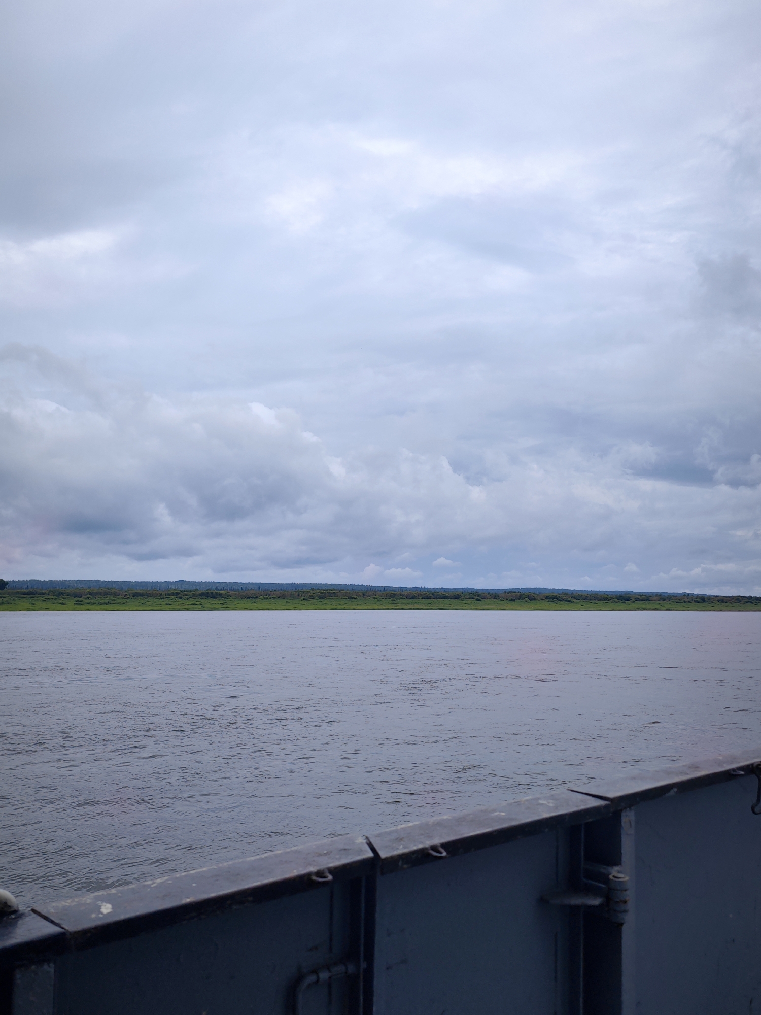 This image captures a wide view of a river under a cloudy sky. In the foreground, the dark railing of a vessel, likely a ferry or barge, is visible. The river's surface is textured with small ripples. Across the river, a dense line of green trees forms the distant shoreline. The sky is filled with a mix of gray and white clouds, suggesting an overcast day. The scene appears to be located in the vicinity of Zotino, Russia, a settlement situated on the Yenisei River. There are no people or specific actions depicted in the image, only the natural environment and a man-made structure.
