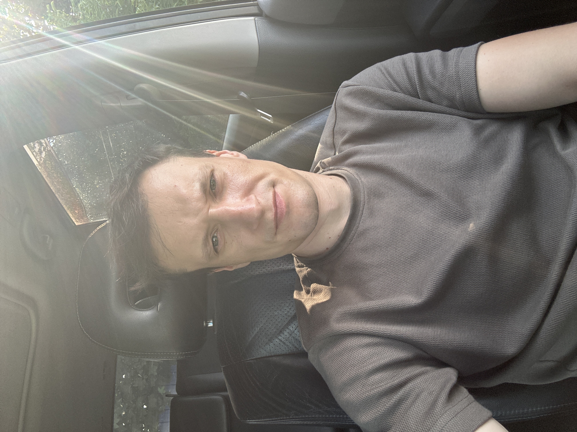A medium shot captures a man with fair skin and dark hair, seated in the driver's seat of a vehicle. He is facing forward, looking directly at the camera with a neutral expression. The man is wearing a short-sleeved, textured brown t-shirt. The interior of the car is visible, including a black leather headrest and seat. Sunlight streams in from the right side of the frame, creating lens flare and casting light across the man's face and shirt. The background outside the car shows foliage, suggesting an outdoor, possibly rural or suburban, location. There are no explicit indicators placing the scene within Shatava, Ukraine; this information is external to the visual data provided.
