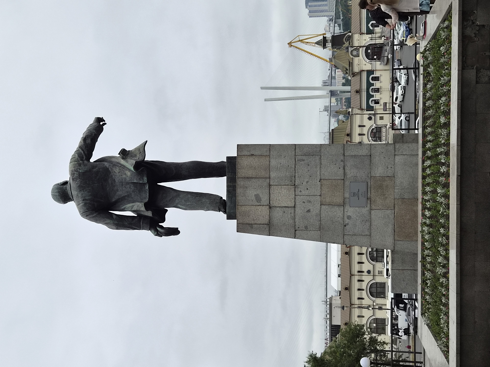 A full shot shows a statue of a man in a suit from behind, standing on a large granite pedestal. The man is holding a hat in his right hand and pointing with his left. In the background, a city skyline is visible, featuring buildings, a bridge, and a crane. The sky is overcast. The statue is located in Vladivostok, Russia, likely at the Nikolai's Triumphal Arch or nearby. People are seen walking in the foreground and background, and cars are parked in a lot. The area appears to be a public space with planters in the foreground.