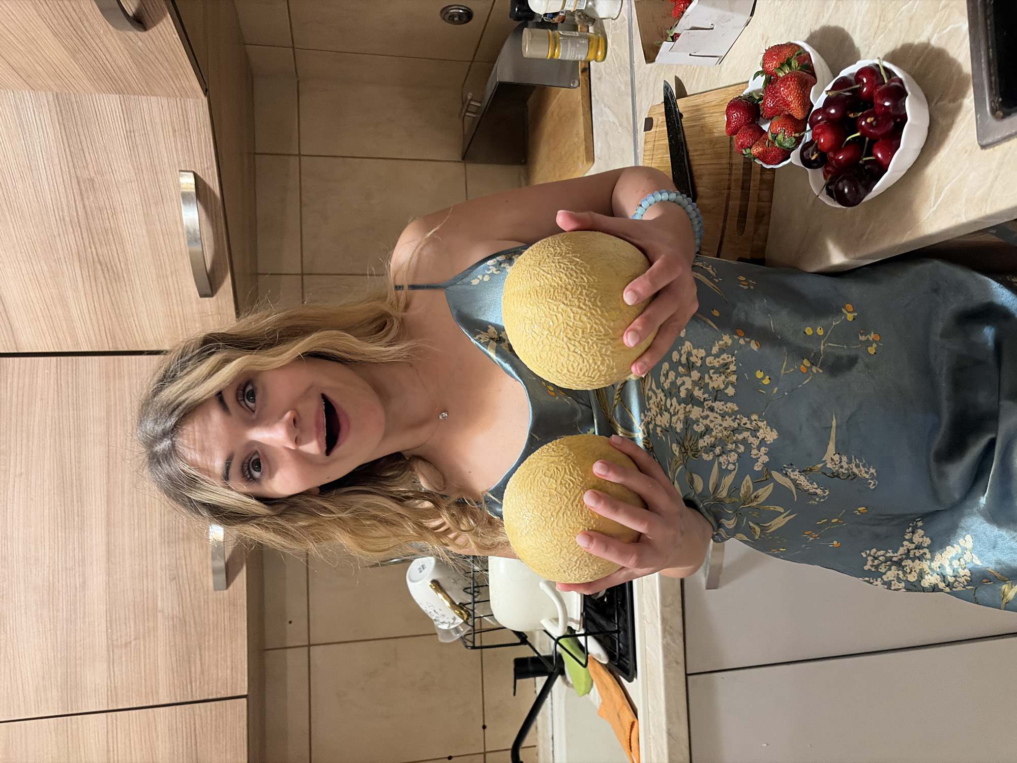 A woman with long blonde hair is holding two cantaloupes in a kitchen. She is wearing a blue floral-patterned dress. In the background, there are two heart-shaped bowls, one filled with strawberries and the other with cherries, on a cutting board. The kitchen has light-colored cabinets and tiled walls. The location is Budva, Montenegro.