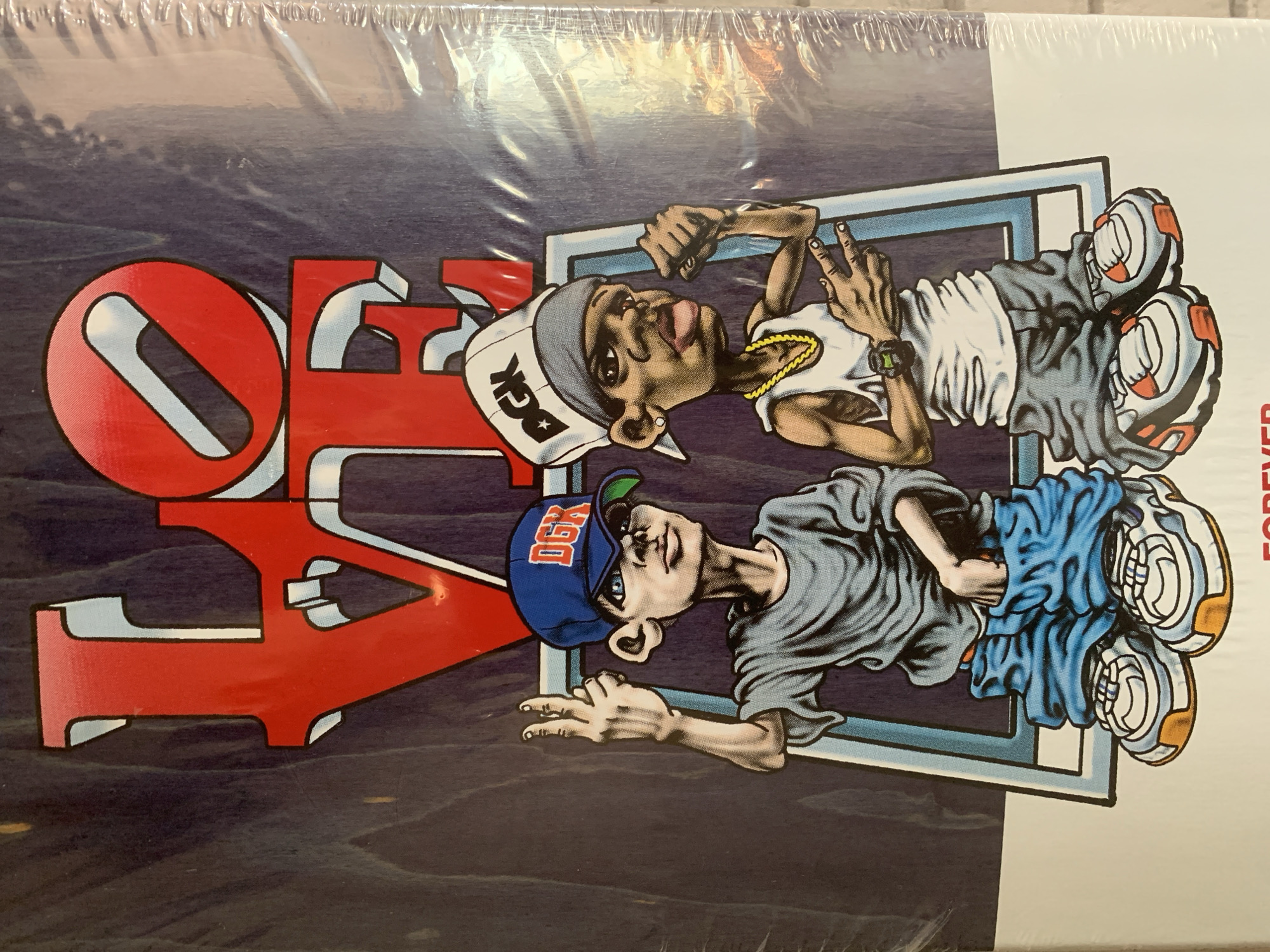 A vibrant, cartoon-style graphic is depicted on what appears to be a product cover, visible through crinkled clear plastic packaging. The main subjects are two stylized male figures positioned within a blue and white rectangular frame.
The figure on the left has darker skin and is wearing a white backward cap with the letters "DCK" (likely a stylized "DGK"), a white tank top, and a gold chain. He holds his left hand up in a gesture that resembles a peace sign or "rock on." The figure on the right has lighter skin, wears a blue backward cap with "DGK," a grey t-shirt, and blue baggy shorts. He is making a two-finger peace sign with his left hand. Notable details include a green digital watch on one figure's wrist and elaborately detailed sneakers on both.
Prominently displayed on the left side, overlapping the figures and background, is large, bold, red stylized text spelling "DGK," rendered with light blue and white outlines for a 3D effect. The background behind the characters and text features a dark, wavy blue pattern. Partially visible along the right edge is vertical red text that reads "FOREVER." The overall aesthetic suggests a connection to skate culture or hip-hop art.