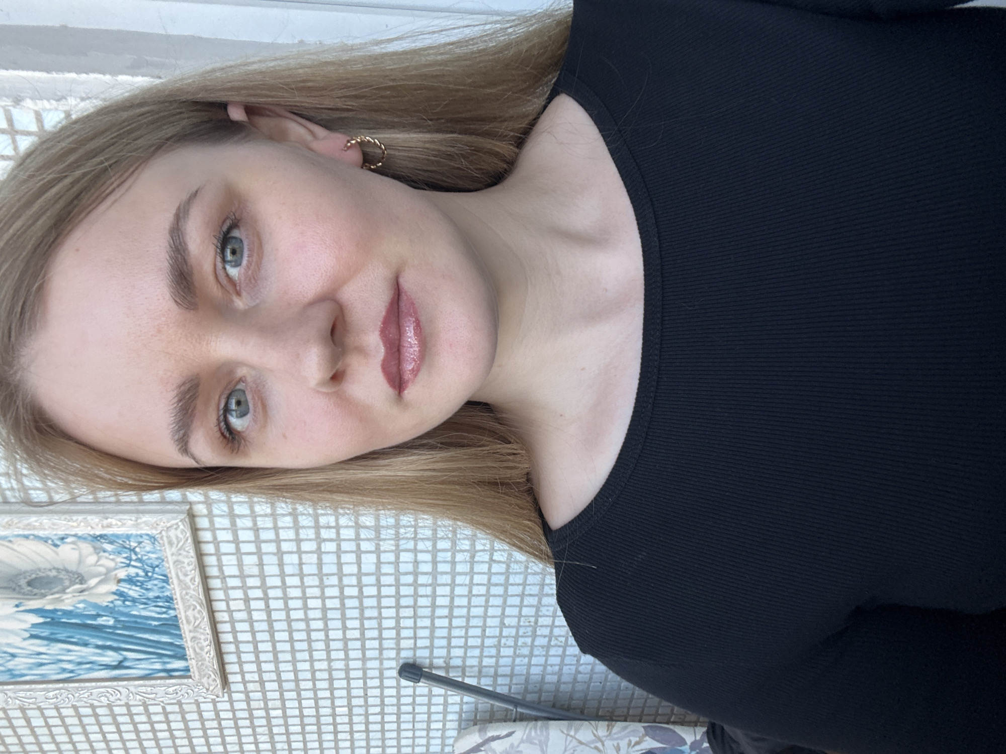 The image depicts a forensic close-up portrait of a single individual, a female, situated in what appears to be an indoor or semi-enclosed setting within the city of Chaikovsky, Russia. The female subject, appearing to be in her 20s or 30s, has light skin, long straight blonde hair worn down, blue eyes, and light brown eyebrows. She is looking directly at the camera with a neutral expression, exhibiting subtle makeup including eyeliner and a rosy-pink, glossy lip product. She wears a black, long-sleeved, ribbed crew-neck top. A small, twisted, gold-colored hoop earring is visible in her right ear. The background consists primarily of a wall covered in small, square, light-colored mosaic tiles. On the upper left of the tiled wall, a framed artwork featuring a white flower against a blue, vertically striped background is displayed in an ornate white frame. To the immediate left of the subject, a slender, dark-colored object with a black cap or handle leans against the tiled wall. A patterned fabric item, possibly upholstery, is partially visible on the far left. A light-colored vertical structure, likely a door or window frame, is visible along the right edge of the image. The lighting is even, suggesting an interior or shaded exterior environment.