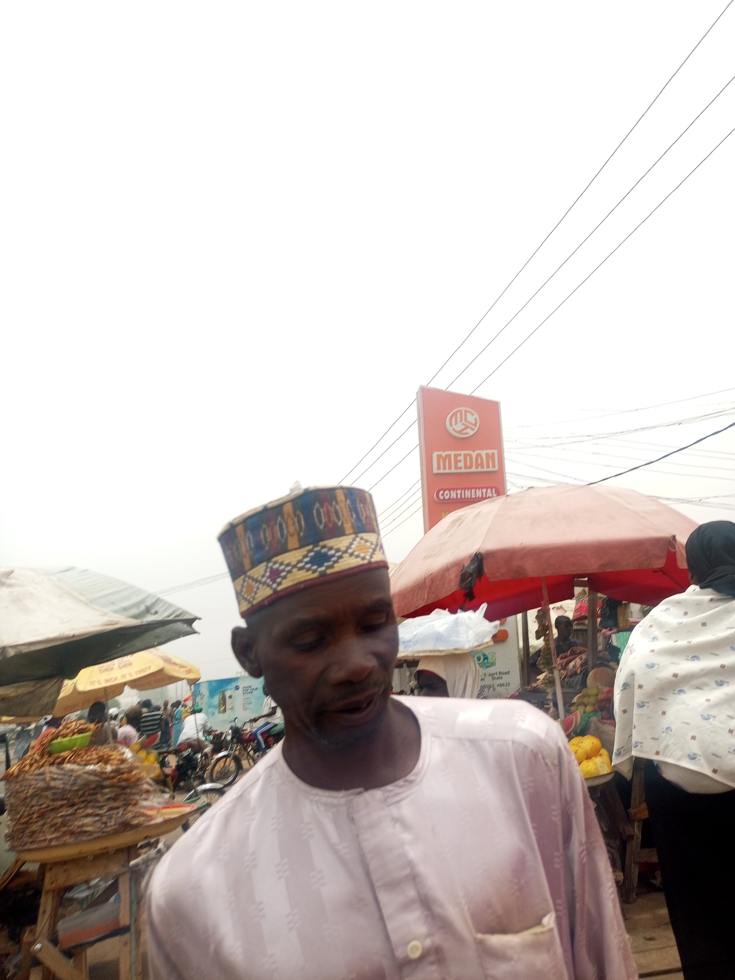 A slightly blurred, eye-level shot captures a vibrant market scene in Tempure, Nigeria, with a man in the foreground. The man, positioned slightly off-center to the left, has dark skin and wears a light-colored, long-sleeved traditional tunic with subtle vertical patterns. His head is adorned with a colorful, patterned cap featuring geometric designs in blue, yellow, red, and white. His gaze is directed downwards and to the right, and his expression is somewhat obscured by motion blur.
Behind him, a bustling open-air market unfolds. To his left, under a light-colored umbrella, is a stall laden with what appears to be a large quantity of dried fish or shrimp stacked on a wooden table. Further back, another yellow umbrella is visible, and the scene is populated by other individuals and motorcycles, suggesting a busy thoroughfare or market alley.
To the right of the man, a person wearing a white, patterned garment and a black head covering is partially visible, facing away from the camera. A red market umbrella canopy dominates the mid-ground, providing shade for another stall where fruits, possibly yellow plantains or mangoes, are displayed.
Prominently displayed above the red canopy is a large, orange advertising sign with white lettering. The sign clearly reads "MEDAN" in a large font, with "CONTINENTAL" written below it in smaller text. Electrical wires crisscross the mostly white, overcast sky above the market. A smaller sign, partially visible beneath the dried fish stall, includes text like "J. Sari Road Shaki" and a phone number starting with "0802".
The overall atmosphere is that of a lively, everyday market environment under a dull, possibly cloudy, daytime sky.