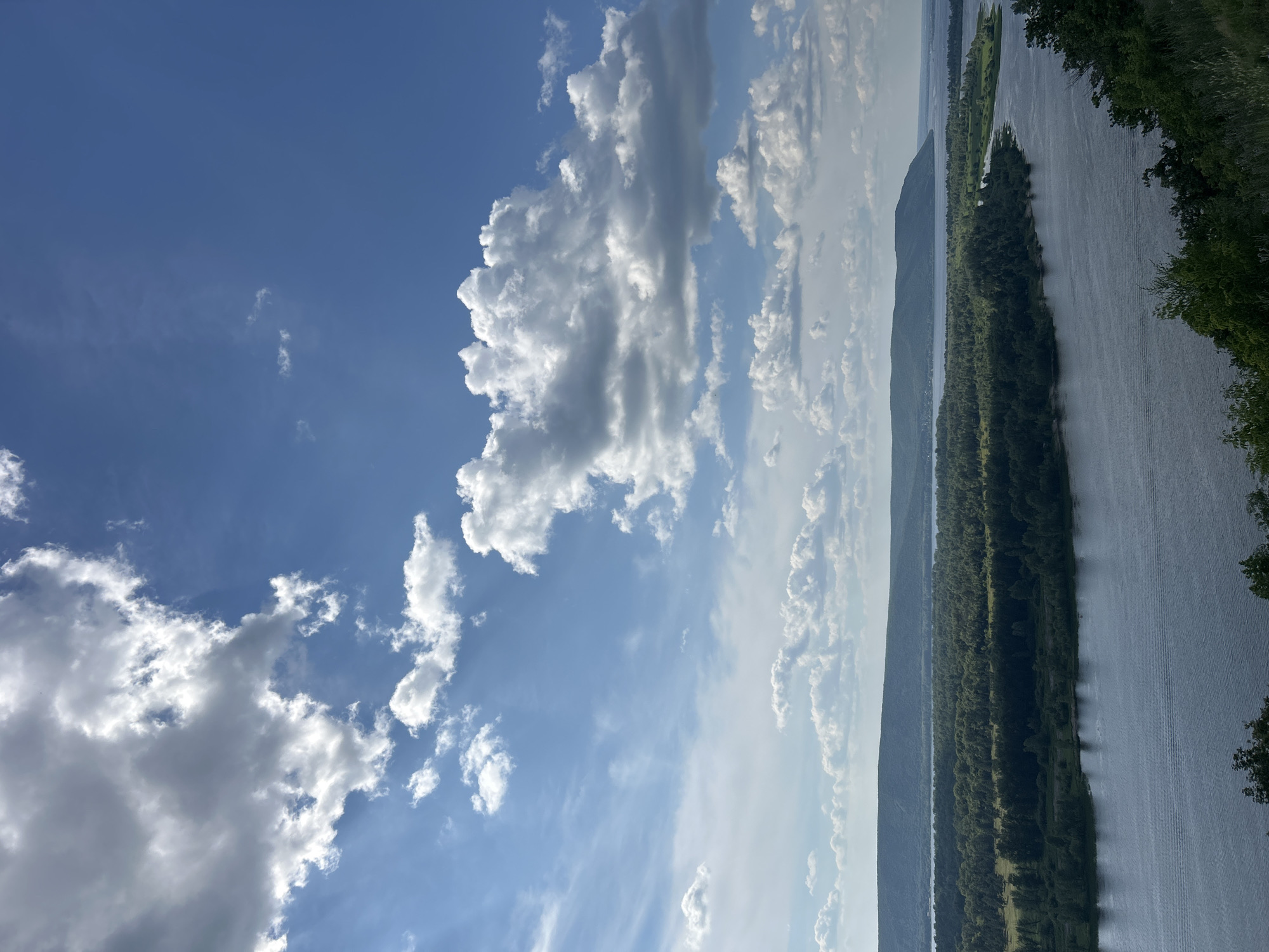 This media file showcases a serene landscape scene under a bright, partly cloudy sky. The primary subjects are a vast body of water, a long, tree-lined island or peninsula, and a distant mountain range. The water, likely a river or lake, reflects the sky and the surrounding greenery, creating a shimmering effect. Dense green trees cover the island, with some lighter green patches that might indicate grassy areas. The distant mountains are rendered in soft blues and grays, fading into the hazy horizon.
The image appears to be taken during the daytime, likely mid-morning or afternoon, given the bright sunlight illuminating the clouds and the landscape. The weather is fair and pleasant, with a mix of fluffy white clouds and thinner, wispy ones against a clear blue sky. The overall mood is one of tranquility and natural beauty.
There is no visible text in the image. The scene, with its wide river, forested islands, and rolling hills, suggests a location in a temperate region with significant natural waterways and woodlands. Based on the provided location context of Novokuybyshevsk, Russia, this scene likely depicts a view of the Volga River or a similar large river system common to the area. The perspective implies the shot was taken from an elevated position, perhaps a hill or a viewpoint overlooking the river.