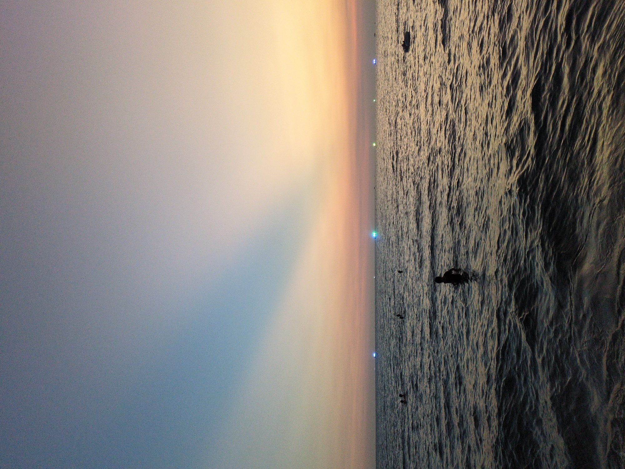 The image depicts a seascape at dusk in Phú Quốc, Vietnam. The sky transitions from blue to orange towards the horizon. The sea is textured with ripples. Lights, possibly from boats or shoreline structures, are visible in the distance. Several figures, possibly swimmers, are in the water.