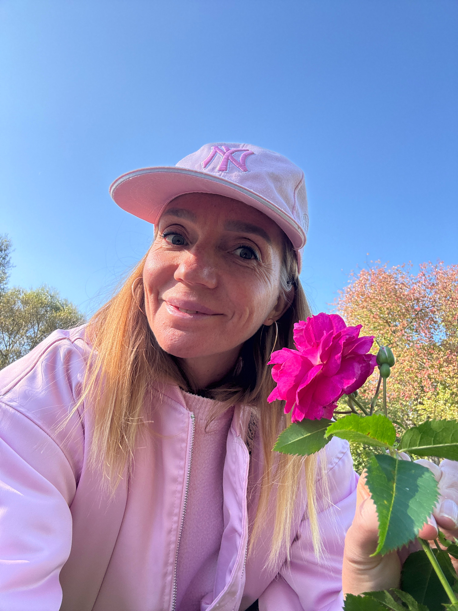 The image shows a woman with blonde hair wearing a pink baseball cap with a pink "NY" logo and a pink jacket. She is holding a vibrant pink rose. The woman is smiling and looking at the camera. Behind her, a tree with pink and green leaves is visible against a clear blue sky, suggesting a sunny day. The scene appears to be outdoors, possibly in a park or garden. The overall impression is cheerful and bright, with the pink theme dominating the woman's attire and the rose. The time of day appears to be daytime due to the bright lighting and clear sky. There are no specific cues indicating Saint Petersburg, Russia, as the location.