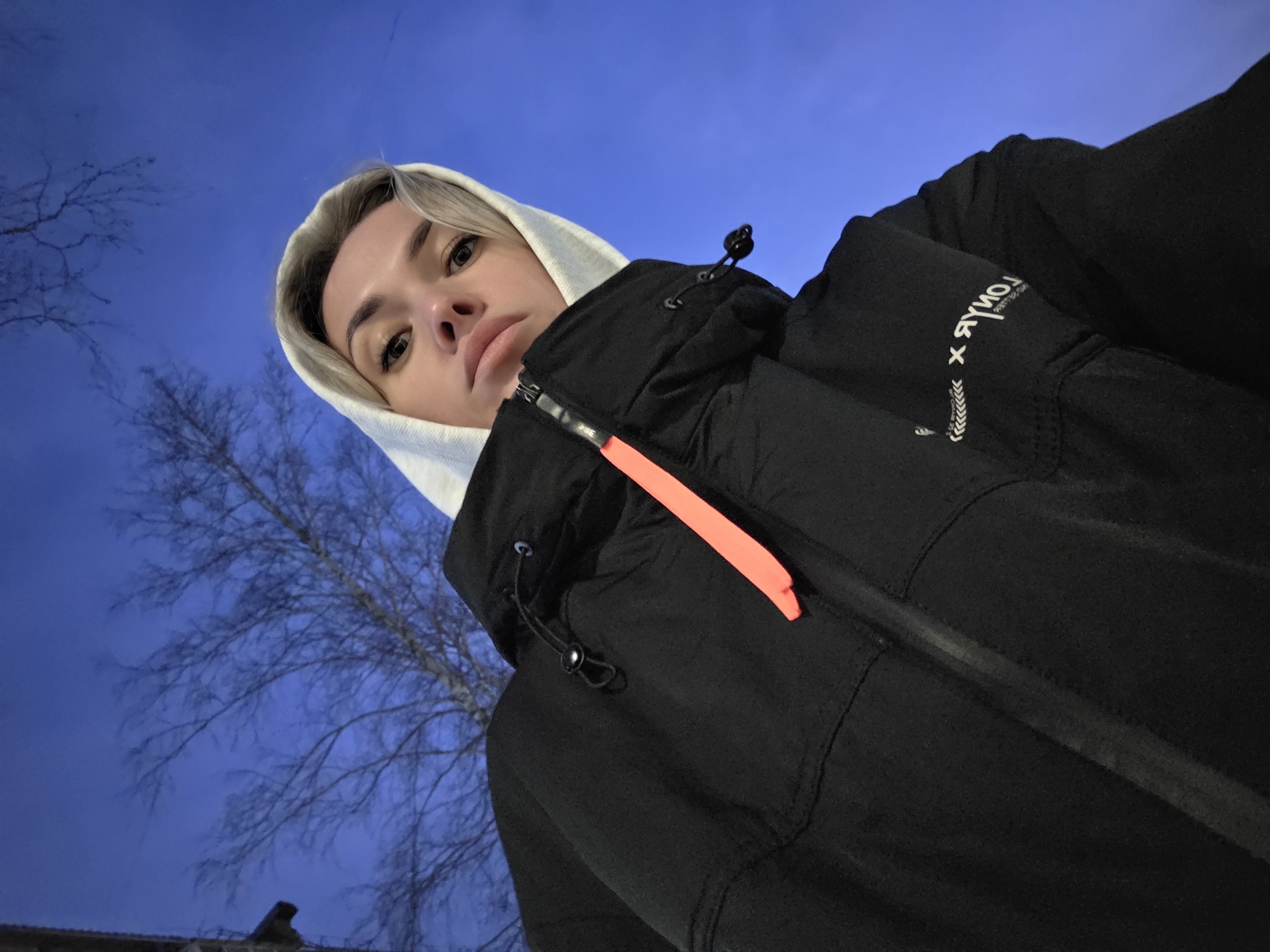 The image is a low-angle selfie of a person with light blonde hair wearing a white hooded sweatshirt under a black jacket. The jacket has a bright orange strip near the zipper. The person's expression is neutral. The background is a blue sky with bare tree branches visible on the left side. The location is the city of Chaikovsky, Russia. The person is likely standing outdoors in an urban environment, possibly near a building, as evidenced by the corner of a roof or wall visible at the bottom.