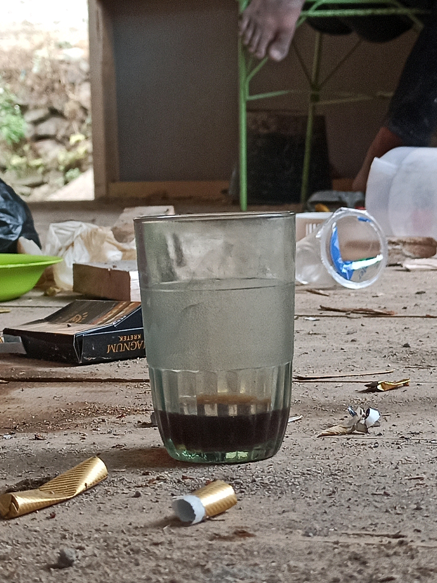 A glass containing a dark liquid sits on a wooden surface. The glass is approximately two-thirds full. Next to the glass is a pack of cigarettes labeled "MAGNUM KRETEK". The surface is cluttered with debris, including discarded cigarette butts and crumpled foil wrappers. In the background, a green metal chair and various plastic containers are visible. The setting appears to be outdoors, possibly in a semi-abandoned or work-in-progress area, given the rough wooden surfaces and scattered materials.