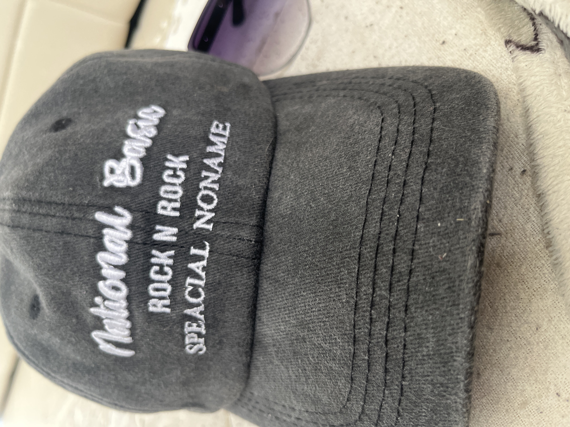 A dark grey baseball cap is displayed in a close-up shot. The cap has white embroidered text on the front. The top line reads "National Basic". The second line reads "ROCK N ROCK". The third line reads "SPEACIAL NONAME". The cap is resting on a light-colored fabric, possibly a towel or blanket. A pair of purple-tinted sunglasses is visible in the upper right corner of the frame, partially obscuring the cap. The background is out of focus and appears to be a light-colored, textured surface. This image was not taken in Chelyabinsk, Russia.