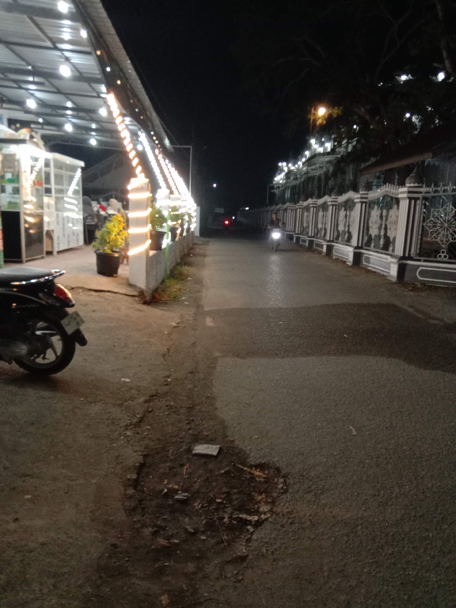 The image depicts a street scene at night in Keudekruenggeukueh, Indonesia. The foreground shows a section of asphalt road with a significant pothole. To the left is a parked motorbike and a brightly lit building with string lights. People can be seen indistinctly inside the building. A series of potted plants line the edge of the building. On the right side of the road is a long, decorative fence bordering a property, with occasional streetlights illuminating the area. A motorbike with its headlight on is moving along the road in the distance. The background is dark, with trees visible.