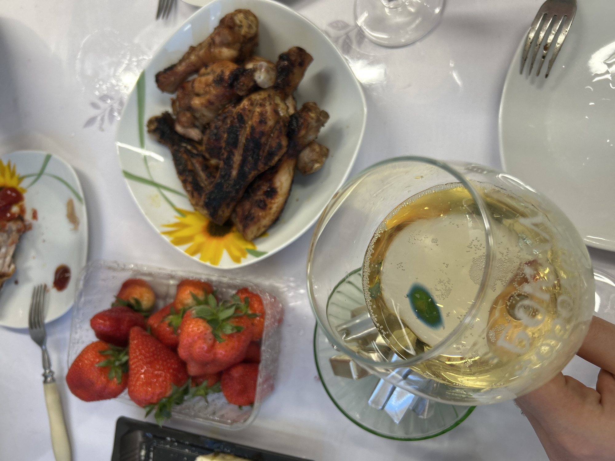 The image presents a table setting with various food items and a drink. A plate of grilled chicken pieces is visible in the upper center, next to a white plate and a fork. A container of strawberries sits on the lower left. A partially filled glass with a yellow liquid, possibly sparkling wine or champagne, is in the lower right corner. Foil-wrapped items are placed under the glass. Other food items are partially visible on a plate in the left part of the image. The background is a white tablecloth with a subtle floral pattern. The photo provides no evidence of location in Bashur, Russia or information about people.