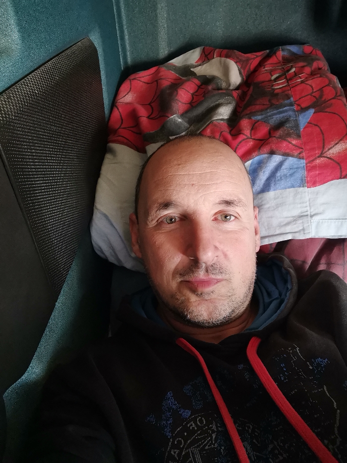 The image displays an adult male, identified as being in Karapelit, Bulgaria, positioned in an enclosed space suggestive of a vehicle sleeper cabin or compact room. The man has a bald crown, short dark hair on the sides, light-colored eyes, and a short beard and mustache. He is wearing a dark-colored hooded sweatshirt with blue hood lining and red drawstrings, featuring a faded graphic on the front. He looks directly forward. To his left, a dark, textured panel and a teal-colored wall are visible. Behind his head and to his right, a light-colored pillow is covered by a red and blue Spider-Man patterned blanket or duvet. A teal-colored surface is also present behind the blanket.