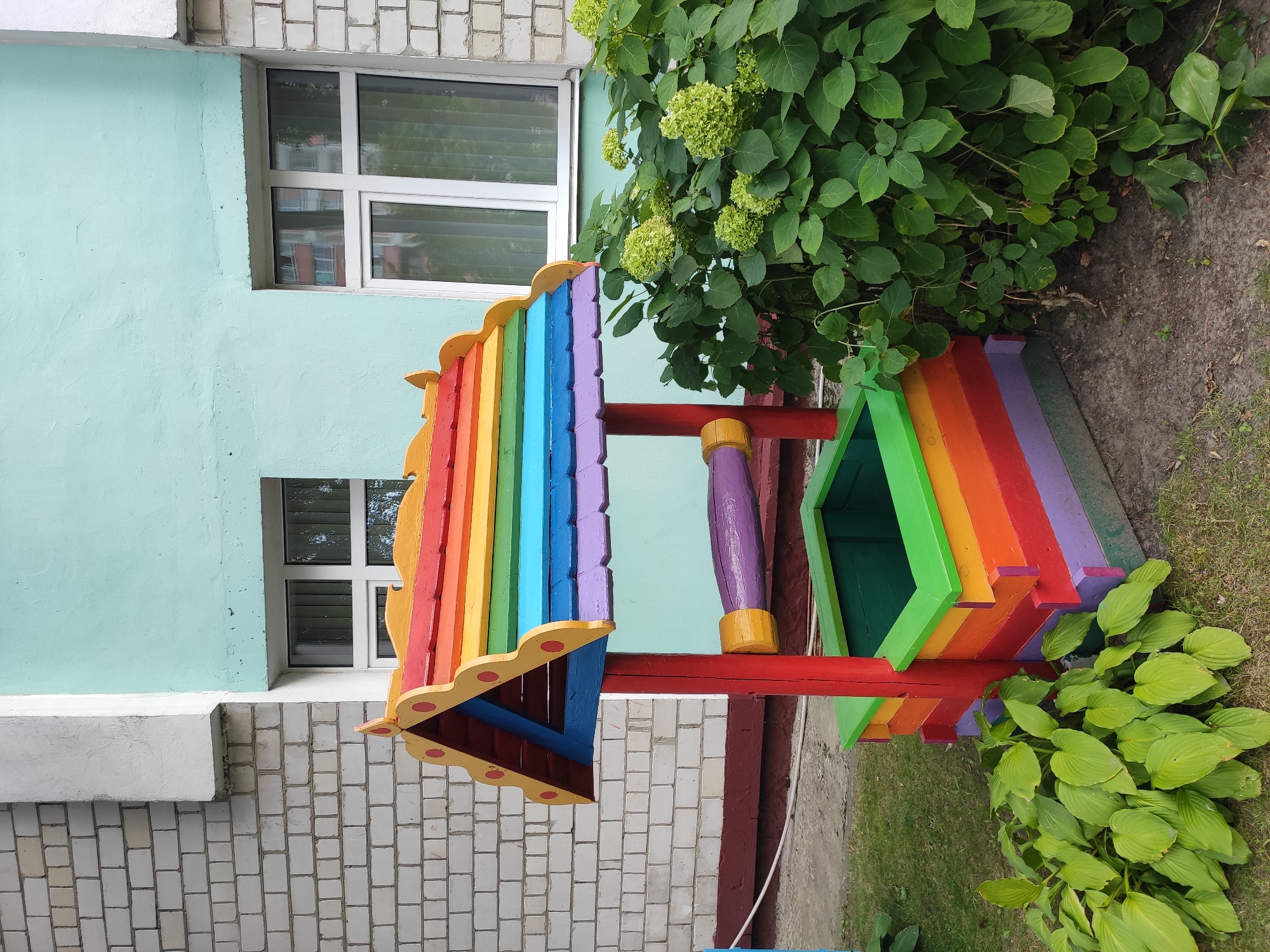 A colorful, rainbow-themed wooden well structure stands in front of a light blue building in Homyel, Belarus. The well is painted with red, orange, yellow, green, blue, and purple stripes, and features a pointed roof. A purple handle is attached to the side of the well. Green plants surround the base of the structure, and a white brick wall of the building is visible to the left, with two windows with white frames.