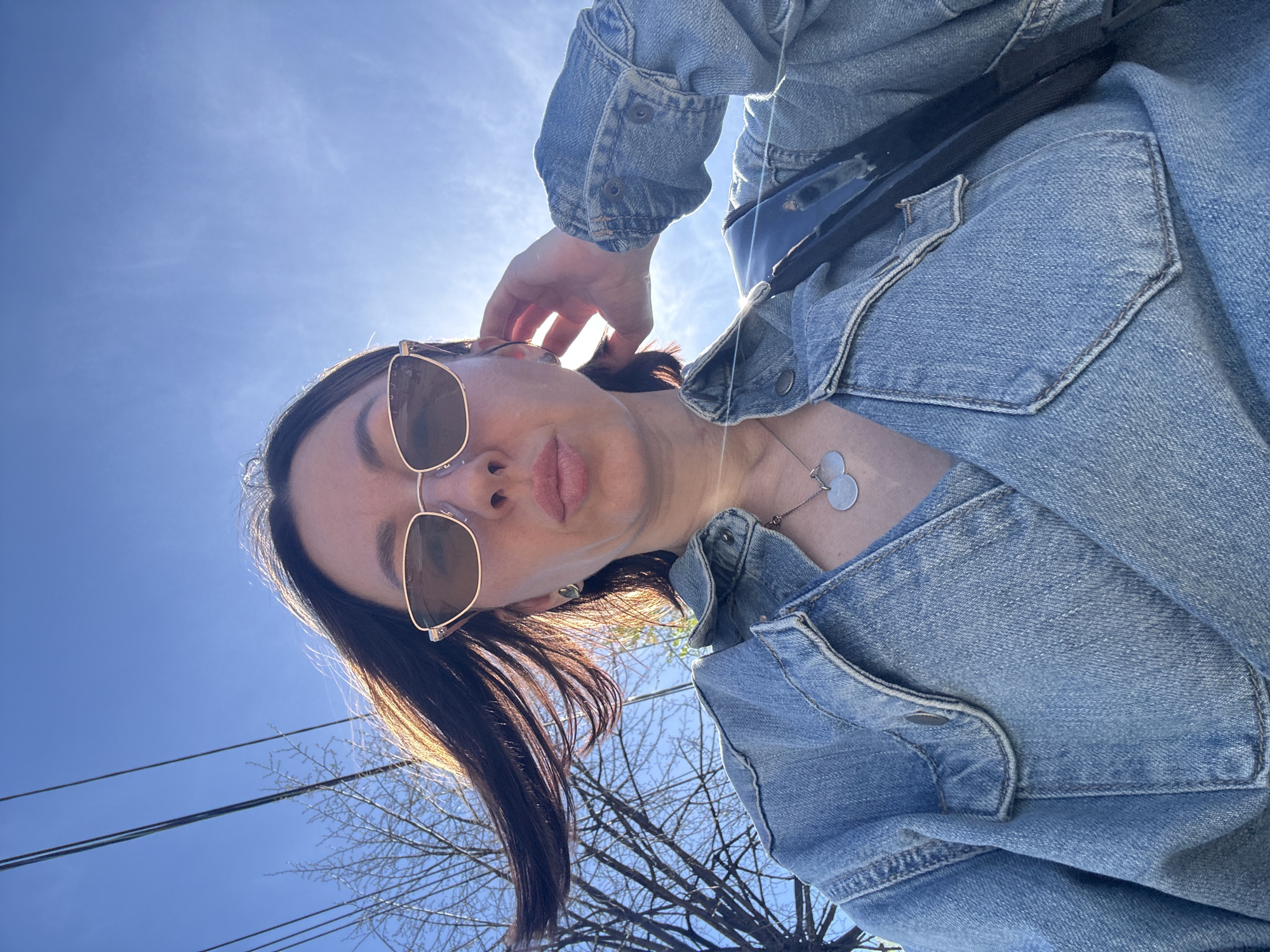 A woman wearing a denim jacket and sunglasses is looking down at the camera. She is wearing a necklace with three circular pendants. In the background, there is a blue sky and bare trees. There are also power lines visible. The image appears to be taken from a low angle.
The image doesn't have enough information to determine that it was taken in Votkinsk, Russia.