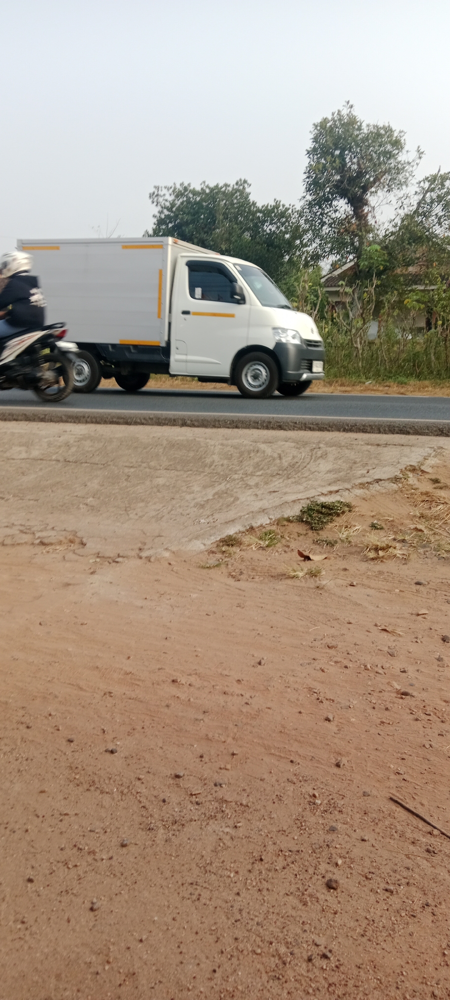 A white box truck with yellow stripes on its sides is driving on an asphalt road. To the left of the truck, a person is riding a motorcycle, also traveling in the same direction. The truck appears to be a delivery vehicle. The background shows a line of trees and vegetation, suggesting a rural or suburban setting. The sky is overcast or hazy. In the foreground, there is a dirt and gravel surface, possibly the shoulder of the road or a path. The scene appears to be captured during daylight. The location is identified as Gondang Rejo, Indonesia.
