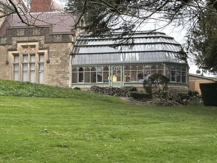 The image displays an exterior view of a stone building connected to a glass conservatory, situated on an upward-sloping green lawn in Malvern Hills, United Kingdom. The main structure, occupying the left, is constructed of light-colored cut stone blocks, featuring multi-paned windows with elaborate stone mullions and decorative carvings above. A section of a brown-tiled roof is visible.
To the right, an attached, multi-faceted glass conservatory features a green metal framework and a segmented glass roof. Numerous transparent window panes form its walls, through which internal illumination is visible. A set of green double doors with a pointed arch design is centered on one of the conservatory's facets. The conservatory rests on a low, rough stone retaining wall, fronted by various small shrubs and plants.
The foreground is dominated by a neatly mown, bright green grass lawn that slopes gently upwards towards the structures. Bare tree branches partially obscure the upper portions of the buildings and an overcast, light grey sky. In the far right background, a section of a tan-colored building with a dark roof and chimney, along with a trimmed hedge, is visible. No individuals or active interactions are present in the scene.