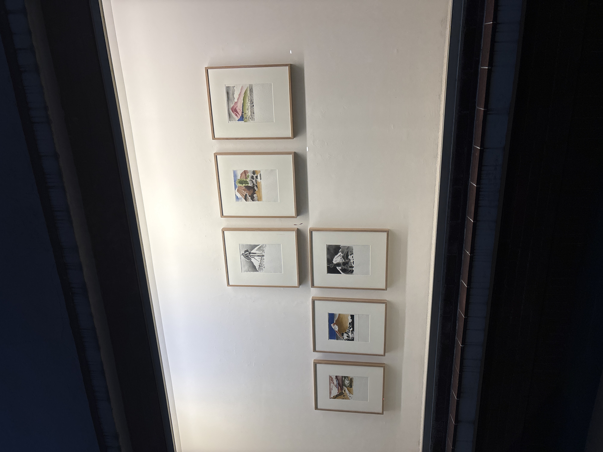 A low-angle shot depicts a vertical arrangement of six framed artworks on a white wall, possibly within an interior space in Mexico City, Mexico. The frames are light brown, and the artworks vary in subject matter and style, including what appears to be watercolor and black-and-white sketches. The lighting is bright, casting a soft glow on the wall and highlighting the frames. The composition is bisected by dark-colored elements on either side, possibly structural components such as beams or architectural details. The perspective suggests the photograph was taken from below, looking upward at the artworks mounted on the wall. No people are visible in the image.