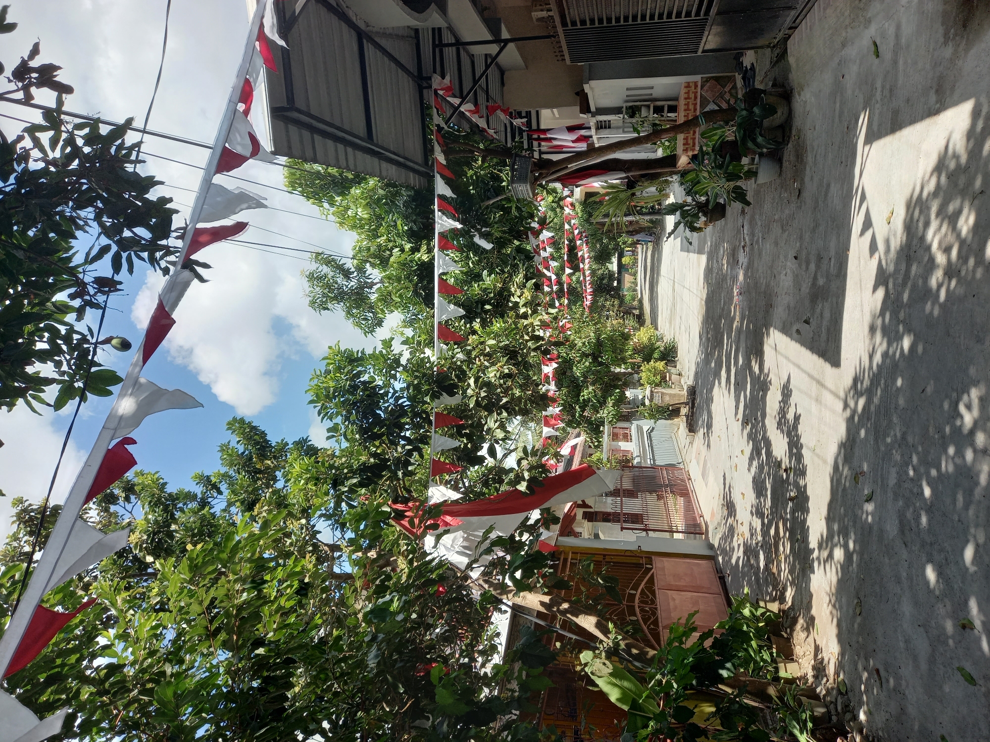 This is a ground-level, eye-level, full shot of a street in Banyumulek, Indonesia, decorated with red and white triangular flags strung across the road. The flags appear to be festive, possibly for a holiday. Trees with lush green leaves line both sides of the street, casting dappled shadows on the concrete pavement. On the left, a closed metal gate of a house is visible, along with potted plants. On the right, houses with awnings and more potted plants are present. In the distance, the street recedes, with a few people visible walking or standing. The sky is blue with scattered white clouds. The overall atmosphere suggests a warm, sunny day in a residential area.