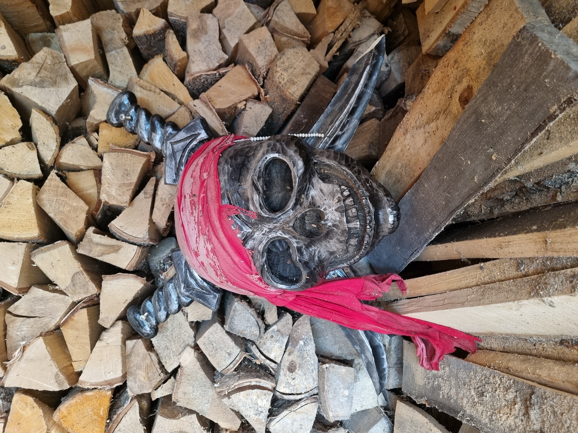 A wooden pile contains firewood and a decorative pirate skull. The skull, made of a dark material, has a red cloth tied around its head and crossed swords behind it. A string of beads adorns the skull's forehead. The image was taken at eye level in an outdoor location in the city of Chaikovsky, Russia.