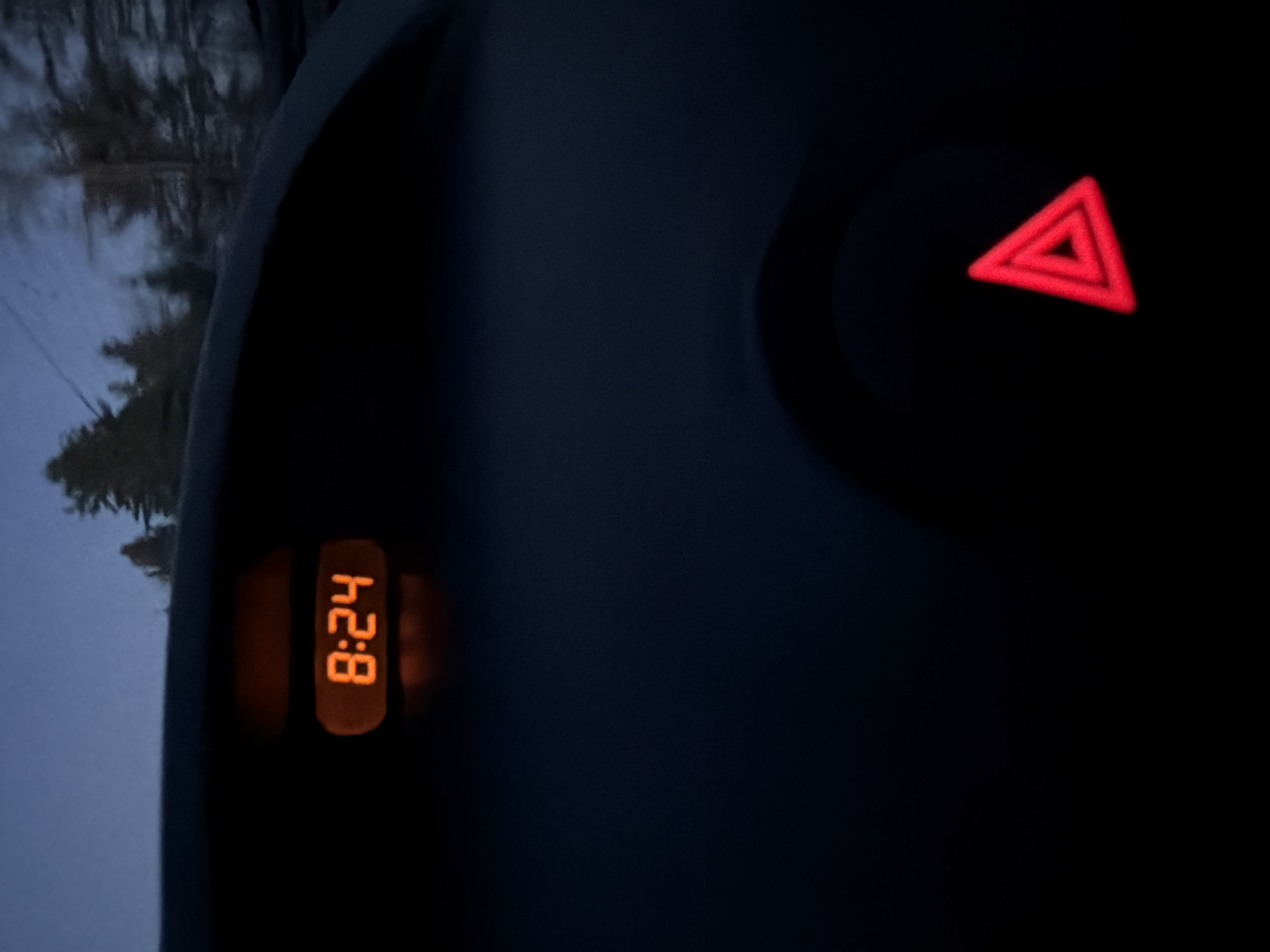 The image shows a car dashboard in darkness. A digital clock displays "8:24" in orange digits. To the right, a red warning light in the shape of a triangle is illuminated. Part of the driver-side window is visible, showing a blurry, tree-lined landscape, suggesting it is early morning or late evening. The location is unknown.