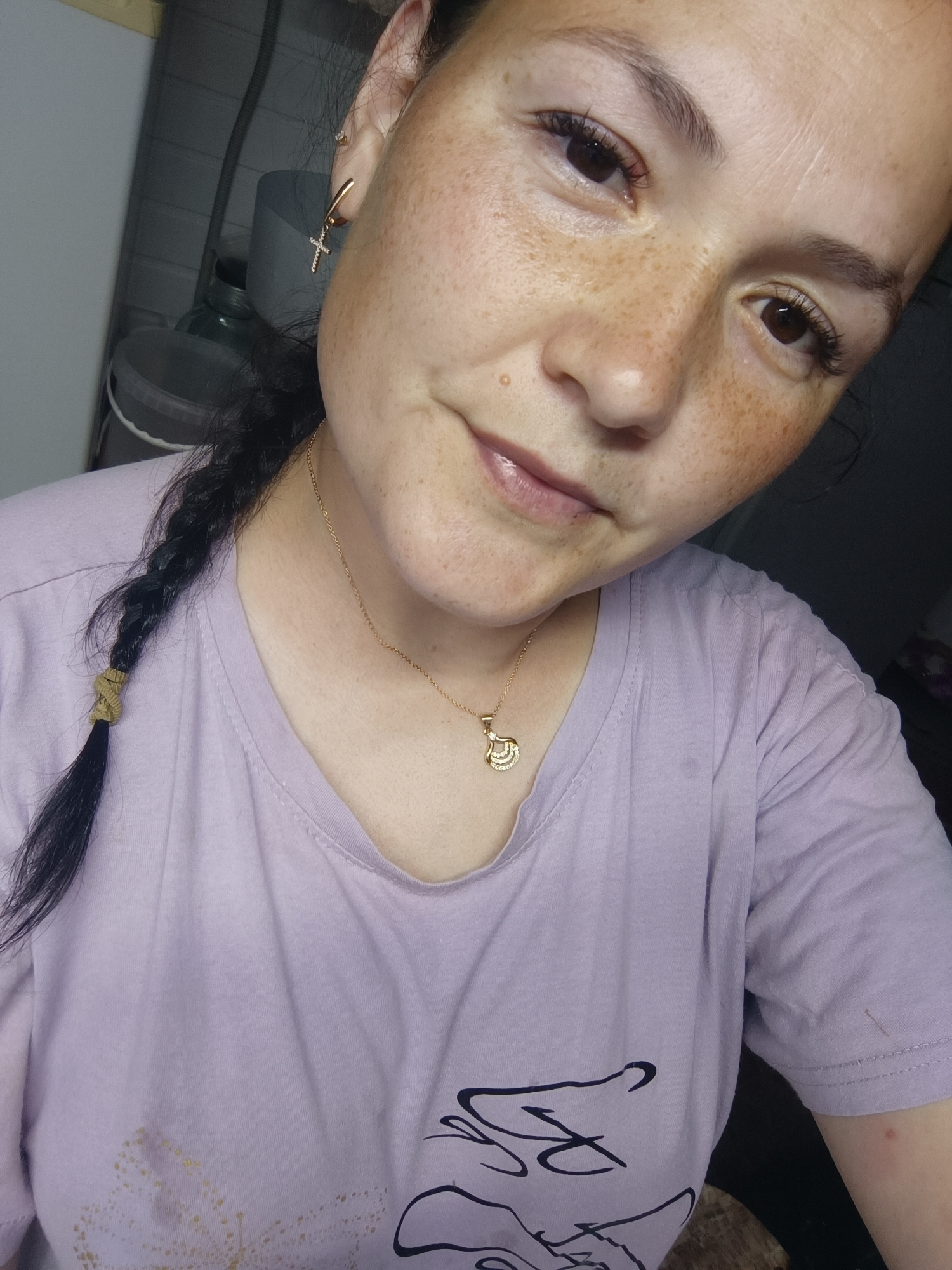 A woman with dark hair in a braid and freckles on her face is shown from the chest up, looking slightly off-camera. She is wearing a light purple t-shirt with a gold pendant necklace. A gold cross earring is visible on her left ear. The background is indistinct but appears to be an indoor setting with some objects that could be containers. The lighting is dim. The image does not contain any information that would allow for the determination of its location in Karsha, Russia.