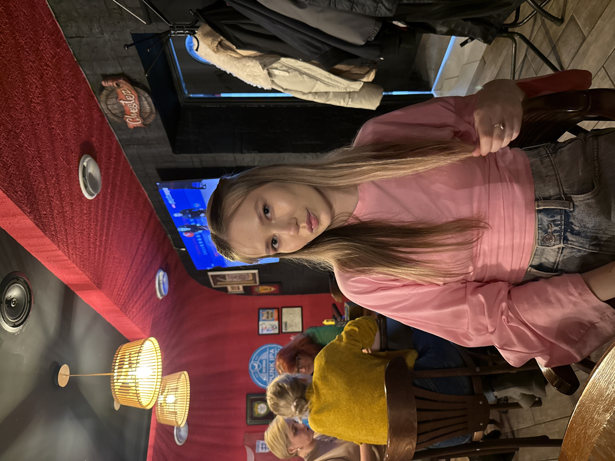The image depicts the interior of Chester Pub, located at Prospekt Pobedy, 25, Chaikovsky, Perm Krai, Russia. A young female is centrally positioned in the foreground, seated at a wooden table, facing the camera. She has long, straight, light-colored hair and wears a pink long-sleeved shirt and light-wash blue jeans. Her right hand is near her side, holding a portion of her hair, displaying a ring on her right ring finger. Her expression is neutral.
In the mid-ground to the left, three other females are seated at a second wooden table. One woman has blonde hair and wears a light-toned top. Another, with light brown hair in a bun, is seen from behind, wearing a bright yellow knitted sweater. The third woman, with red hair, wears a green top and is seen from a partial rear angle.
The environment features a dark ceiling with a red textured fabric running along the wall-ceiling joint on the left. Two wooden slat-style pendant lamps emit warm light, accompanied by recessed ceiling lights and a ventilation grille. The wall behind the background individuals is red, adorned with several framed pictures and a blue circular sign that reads "BREWERY PUNK IPA." To the right, a darker, textured wall section displays a wooden barrel-shaped sign with "Chester" in red script. A flat-screen television showing blue and white graphics is mounted on the wall above the background individuals. A black metal coat rack holding several jackets, including a cream-colored puffer jacket with a faux shearling collar, stands to the right. The floor in the lower right exhibits a tiled pattern.