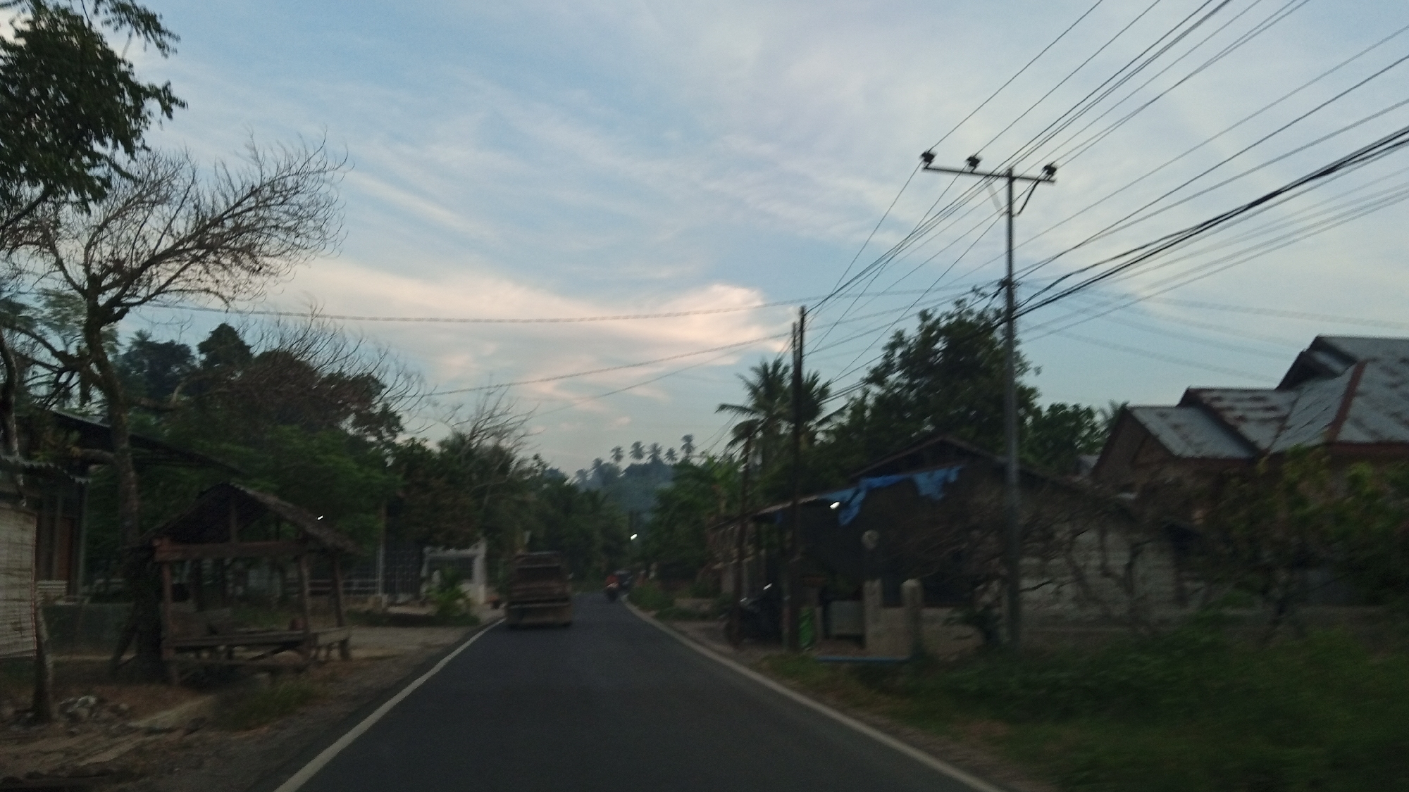 The image depicts a rural road in Luengbaro, Indonesia, during what appears to be dusk or early morning. The sky is a soft blue with wispy clouds. A paved road runs through the center of the frame, with a white line marking the edge. On the left side of the road, there is a small, open-air wooden shelter with a thatched roof, and a bare tree with intricate branches. Further down the road, on the left, are buildings with wooden and brick construction, and dense foliage. On the right side of the road, there are houses with corrugated metal roofs and some areas covered with blue tarps. A utility pole with multiple power lines stands tall on the right, casting shadows. In the distance, a truck is visible on the road, and a motorcycle can be seen further ahead. The landscape is characterized by lush green trees, some of which are palm trees, and a gentle incline of hills in the background. The overall impression is one of a quiet, developing rural community.