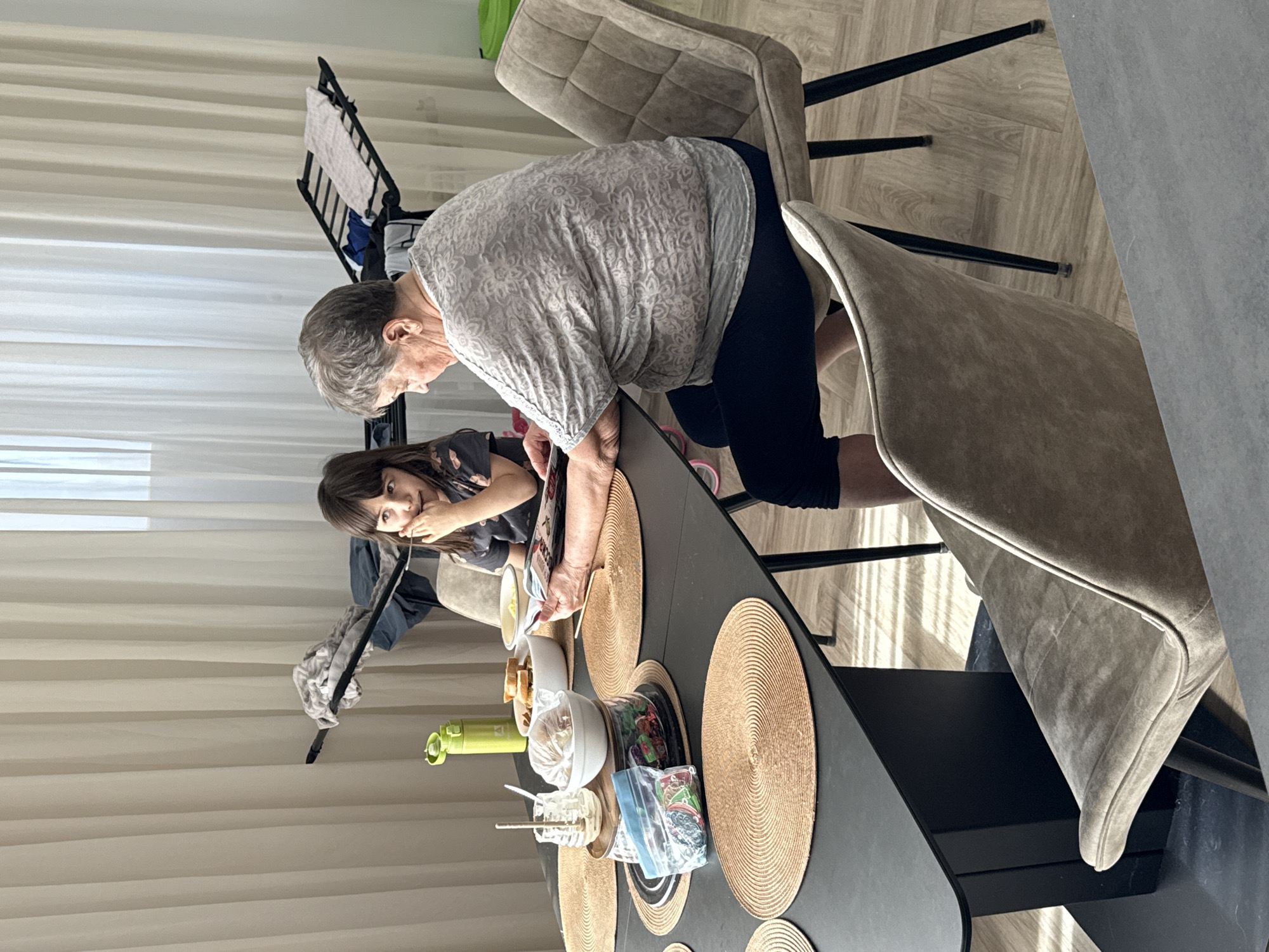 A woman and a girl are at a table. The woman, with short gray hair and wearing a white patterned shirt and black pants, leans over the table, holding a magazine. The girl, with long brown hair, sits beside her, looking at the camera and resting her head on her hand. A black table in front of them is set with woven placemats and various items including containers, a water bottle, and food. Two beige chairs are visible, one of which the woman sits on. The background shows a white curtain and a music stand. This scene is indoors. The location cannot be determined as Krasnoyarsk, Russia.