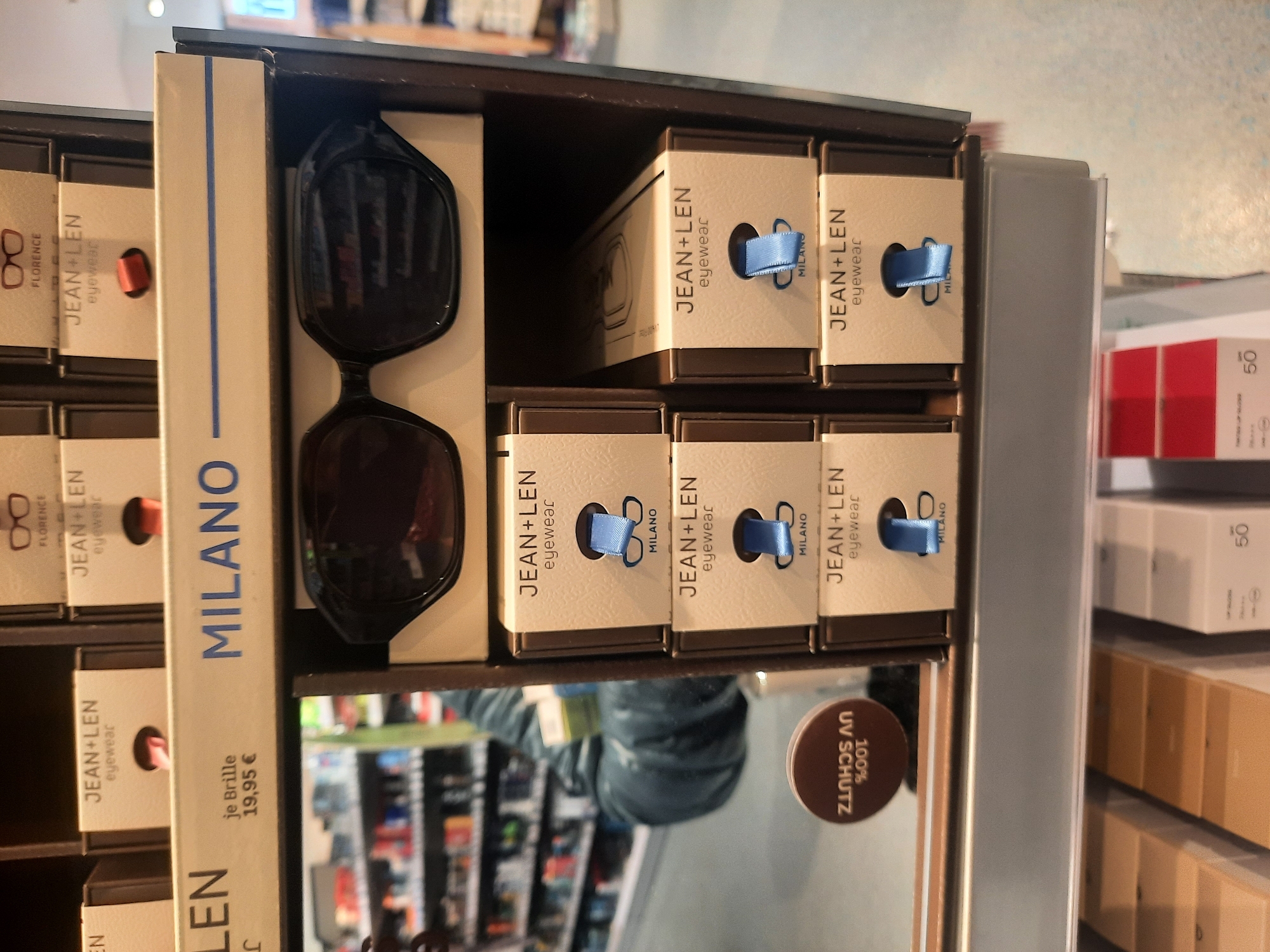 This is a medium shot of a retail display featuring sunglasses. The display is organized vertically, with multiple rows of boxed eyewear. The primary focus is on a pair of dark, large-framed sunglasses positioned prominently on the left side of the image. These sunglasses appear to be resting on a beige box with blue text that reads "MILANO."
To the right of the sunglasses, several smaller beige boxes are stacked. Each of these boxes features blue text that says "JEAN+LEN eyewear" and some have small blue ribbons or bows visible. Some of these boxes also show minimalist line drawings of eyeglasses. Further to the right, a gray vertical plastic divider separates this display from another section. This second section contains several red and beige boxes, some with visible numbers like "50" and text.
A round brown sticker with white text, reading "100% UV SCHUTZ," is partially visible in the lower right quadrant of the image, indicating a feature of the eyewear and suggesting a German-speaking context for "Schutz" (protection). The background is blurred, but suggests a retail environment with shelves stocked with various items, possibly clothing or accessories. The lighting appears to be artificial, consistent with an indoor store setting. There are no people visible in the image. The overall impression is a clean and organized product display.