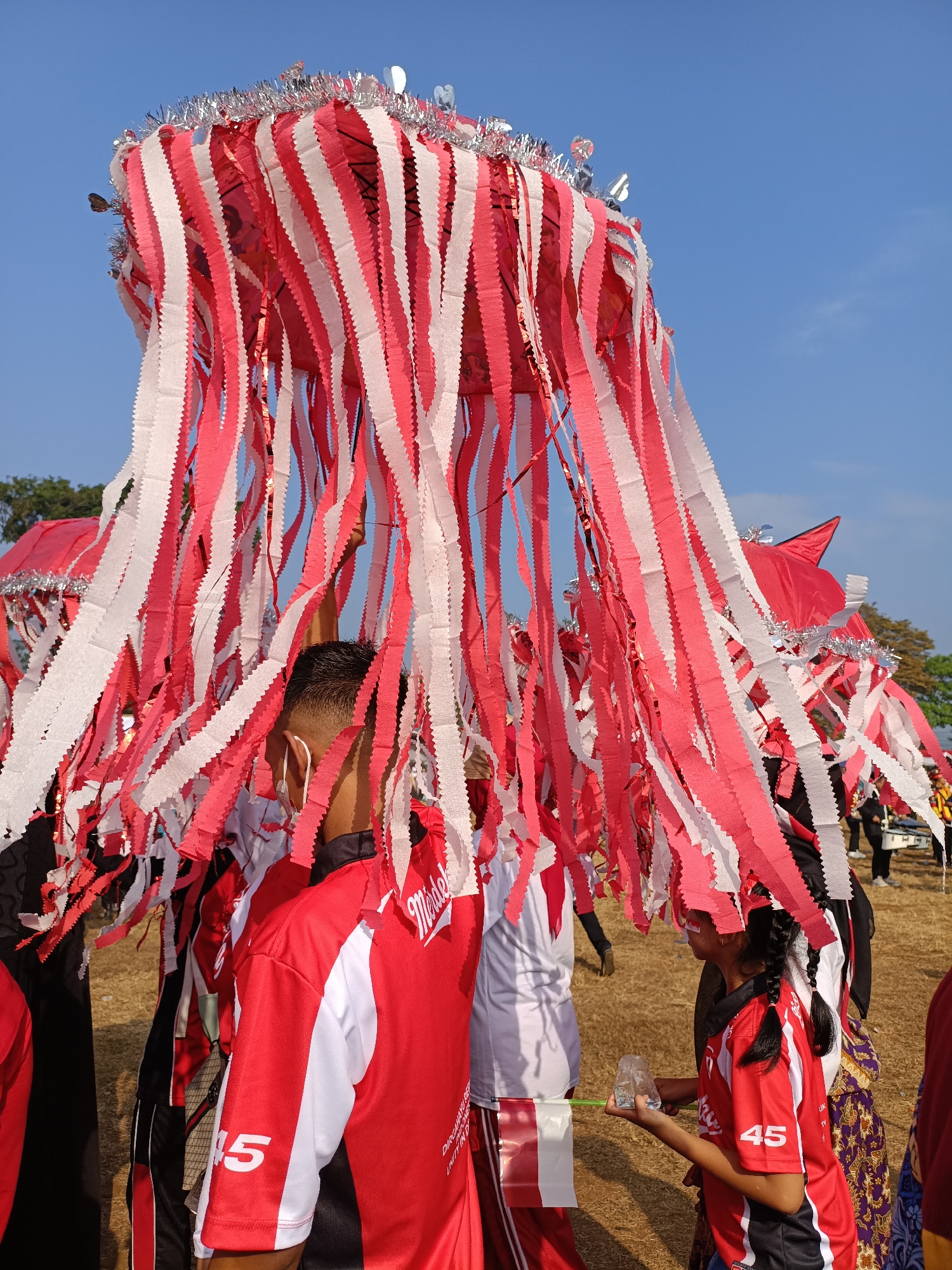 In Cibodas, Indonesia, under a clear blue sky, a group of people are participating in an outdoor event. The foreground is dominated by large, ornate decorations made of alternating red and white paper streamers, adorned with silver tinsel at the top. These streamers hang down, obscuring much of the people beneath.
Several individuals are visible, most wearing matching red and white athletic shirts. One person in the foreground, viewed from the back, has short dark hair and is wearing a white face mask. Another individual, a young girl with braided dark hair, is also in the foreground, holding a small clear plastic container and a green stick. She is wearing a red shirt with the number "45" on the sleeve. In the background, other participants in similar attire are visible, along with what appears to be a marching band drum. The ground is covered with dry, light brown grass. The scene suggests a festive occasion or parade.
