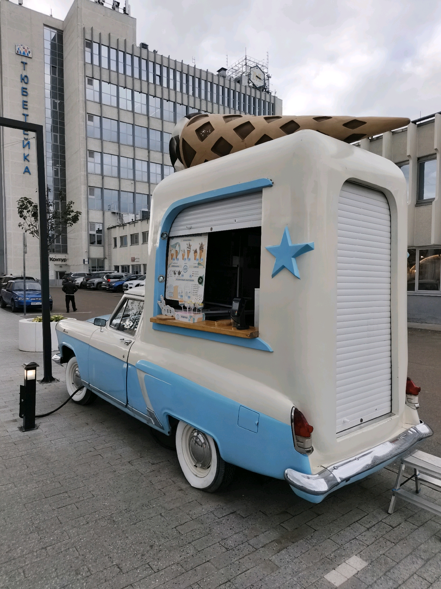 The image shows a vintage ice cream truck, painted in white and light blue, parked on a paved area in front of a modern office building. The truck is a modified GAZ-21 Volga station wagon, with an extended cabin designed to serve ice cream. A large, waffle cone-shaped sculpture is mounted on the roof of the truck. A blue star emblem is affixed to the side of the serving window. Inside the serving window, a menu board displays various ice cream options. A person in a black jacket is walking away from the camera in the background, towards a parking lot filled with cars. The office building features a sign with Cyrillic lettering. The sky is overcast. The overall scene suggests an urban environment, likely a commercial district in Kazan, Russia, given the Cyrillic signage and architectural style.