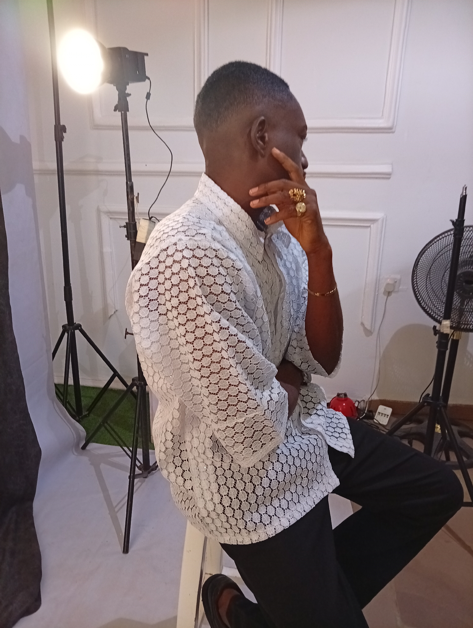 A man sits on a white stool. He is wearing a white lace shirt and black pants. He is wearing multiple gold rings on his right hand and a gold bracelet. His right hand is raised to his face. Behind him is a white wall with a raised square pattern. To his left is a photography light on a tripod. To his right is a black fan on a tripod. There is a white backdrop on the floor, with a patch of green behind the light stand. A red object is visible behind the man, near the fan.