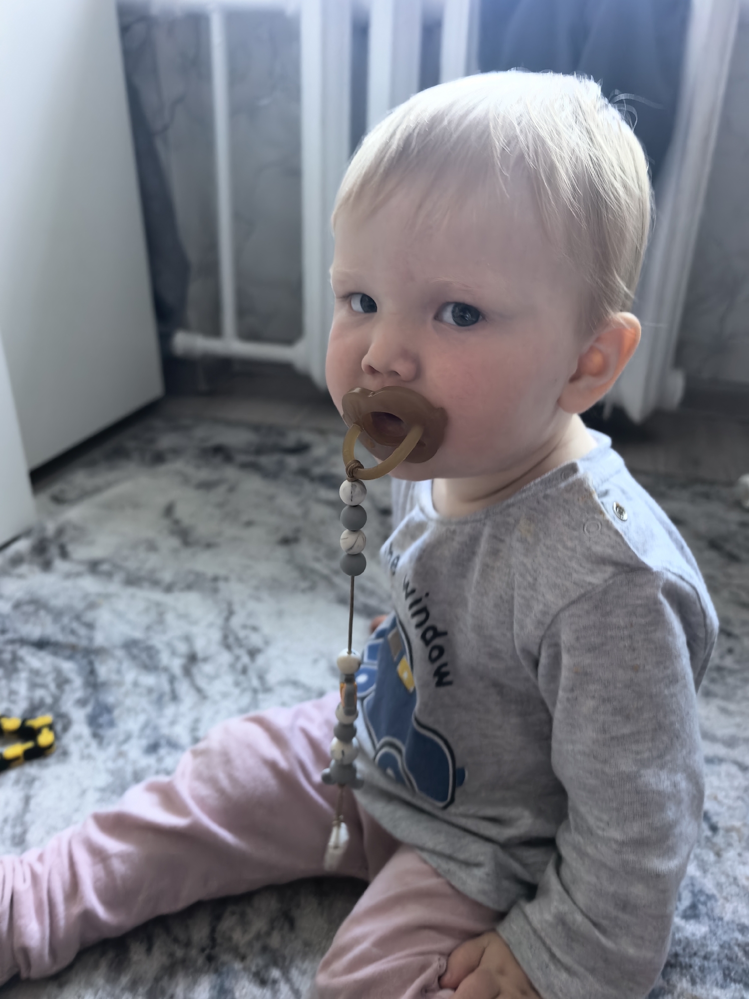 A toddler with blonde hair and blue eyes is sitting on a textured rug in a room, possibly a home. The child is wearing a gray long-sleeved shirt with a blue graphic and pink pants, and has a pacifier in their mouth attached to a beaded clip. The background shows a blurred white and gray wall or furniture. There is a yellow and black toy car on the rug to the left of the child. The scene is indoors and does not provide any information about the city of Sterlitamak, Russia.