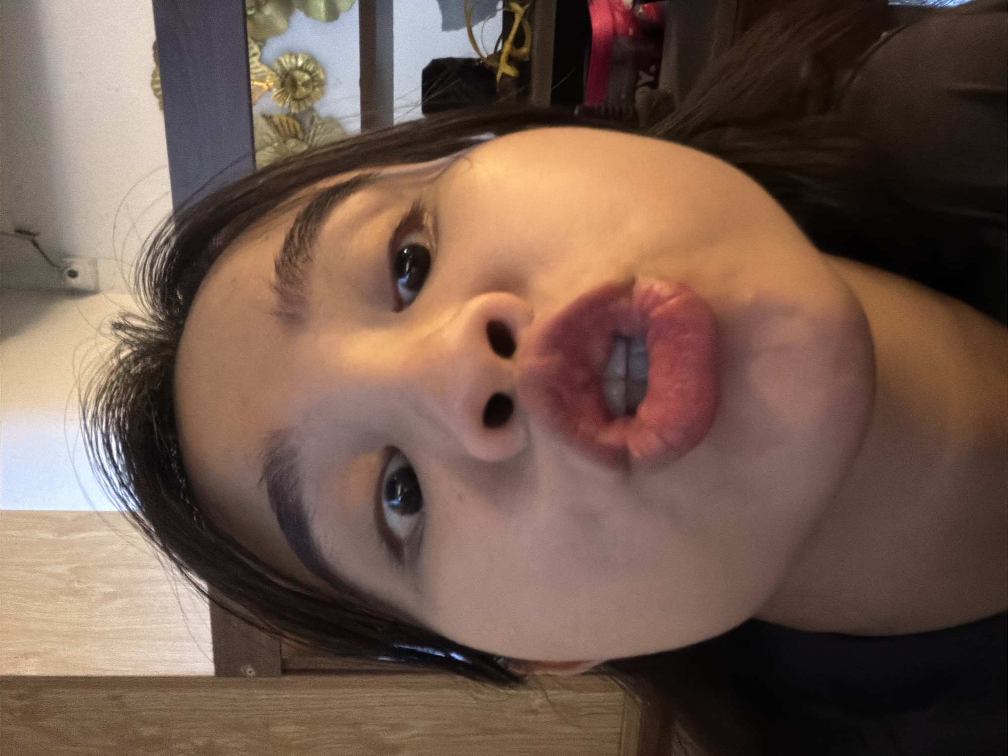 A close-up, rotated portrait captures a young person, likely female, posing playfully indoors in Đà Nẵng, Vietnam. The subject's head is tilted significantly, suggesting the photo was taken from a low or unconventional angle. She has dark hair, dark eyes, and a fair complexion. Her lips are distinctly pursed in a "fish face" or "duck face" expression, revealing the texture of her lips and a glimpse of her teeth. Her right eye is wide open and looking upwards, while her left eye is partially obscured by her forehead. Dark, well-defined eyebrows frame her eyes.
The background indicates an indoor setting. To the upper left, a plain white wall features a small white electrical outlet. Behind the subject's head, in the upper center and right, are a dark vertical structure (possibly a door frame or furniture) and an ornate golden decorative element, potentially part of a mirror or wall art. A wooden surface, appearing as a table or floor, occupies the bottom portion of the frame, extending beneath her head. The lighting is ambient indoor light. The image conveys a casual, lighthearted, and playful moment. No text is visible.