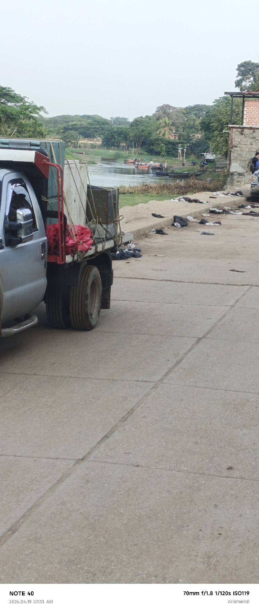 The image, taken in Arismendi, Venezuela on April 19, 2026, at 7:33 AM, shows a partially visible silver pickup truck parked on a paved surface with visible seams. The truck bed contains a metal cage-like structure with a white panel and red bundles. The background reveals a body of water with boats, a shoreline with scattered dark debris, and dense green vegetation. A building is visible on the right side of the frame. People are observed near the building and the shoreline. The image was captured with a 70mm lens at f/1.8, 1/120s exposure, and ISO 119.
