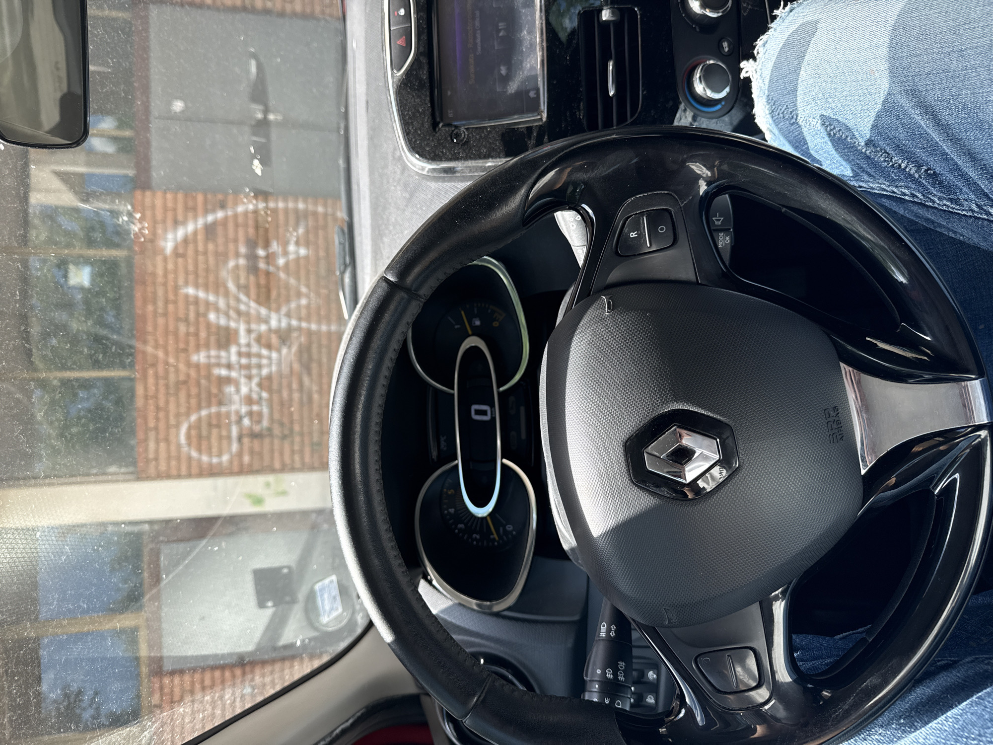 The image shows the interior of a car, with the steering wheel and dashboard prominently featured. The steering wheel has a Renault logo. A person wearing blue jeans is partially visible in the upper right corner. Through the windshield, a building with brick walls and graffiti is visible. The location is Sliven, Bulgaria, as indicated by the graffiti on the building visible through the car's windshield.