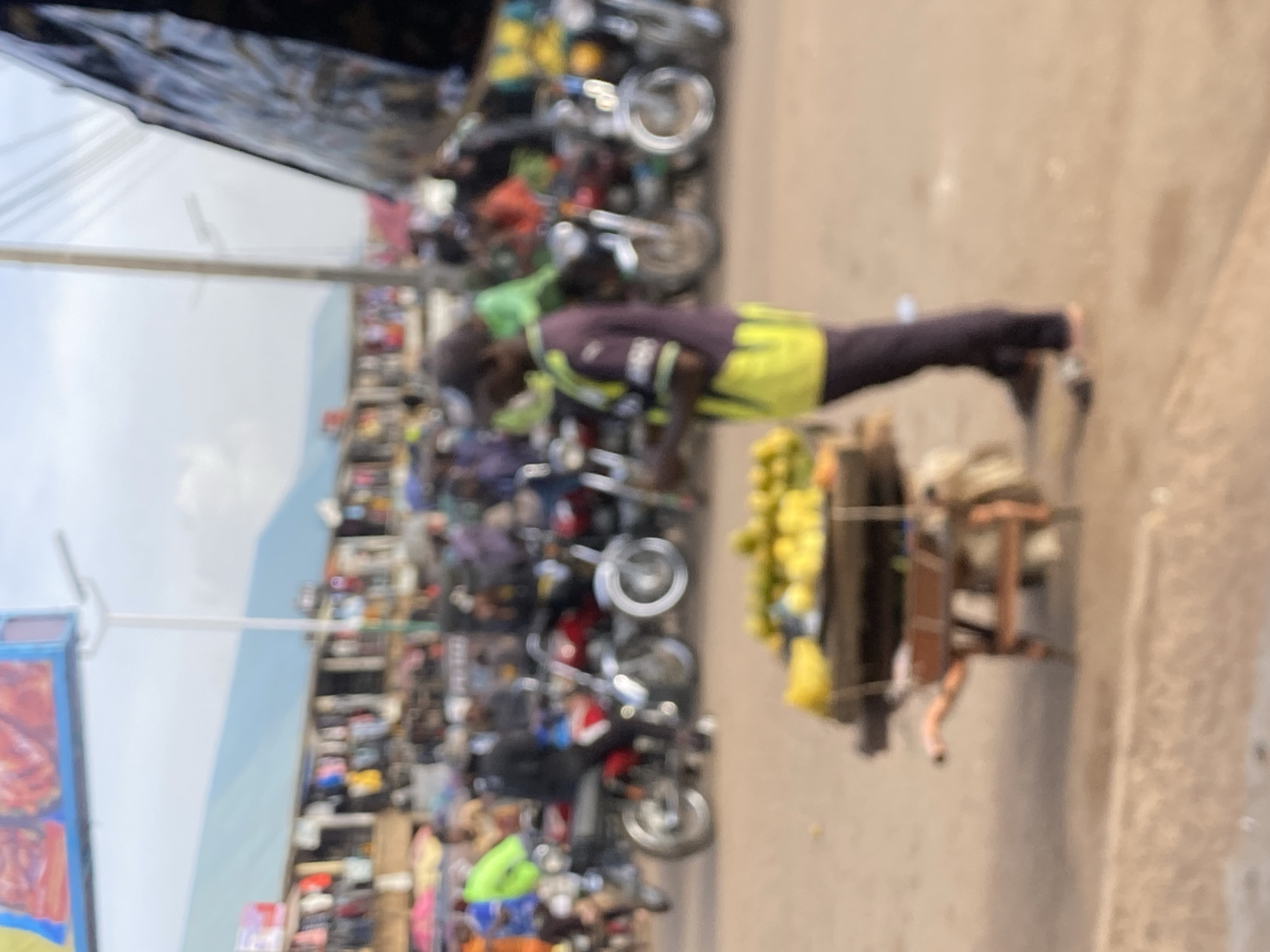 The image depicts a man in Bukuru, Nigeria, pushing a handcart loaded with yellow fruits, likely mangoes or oranges. He is wearing a dark-colored long-sleeved shirt and light-colored shorts, potentially neon yellow. The scene is along a road or street, likely a market area, based on the blurred background of parked motorcycles and the presence of what appears to be a sign or banner on the left. The environment appears crowded, suggesting a busy marketplace atmosphere. The focus is on the man and his fruit-laden cart. The photo is somewhat blurry, making it difficult to discern fine details.