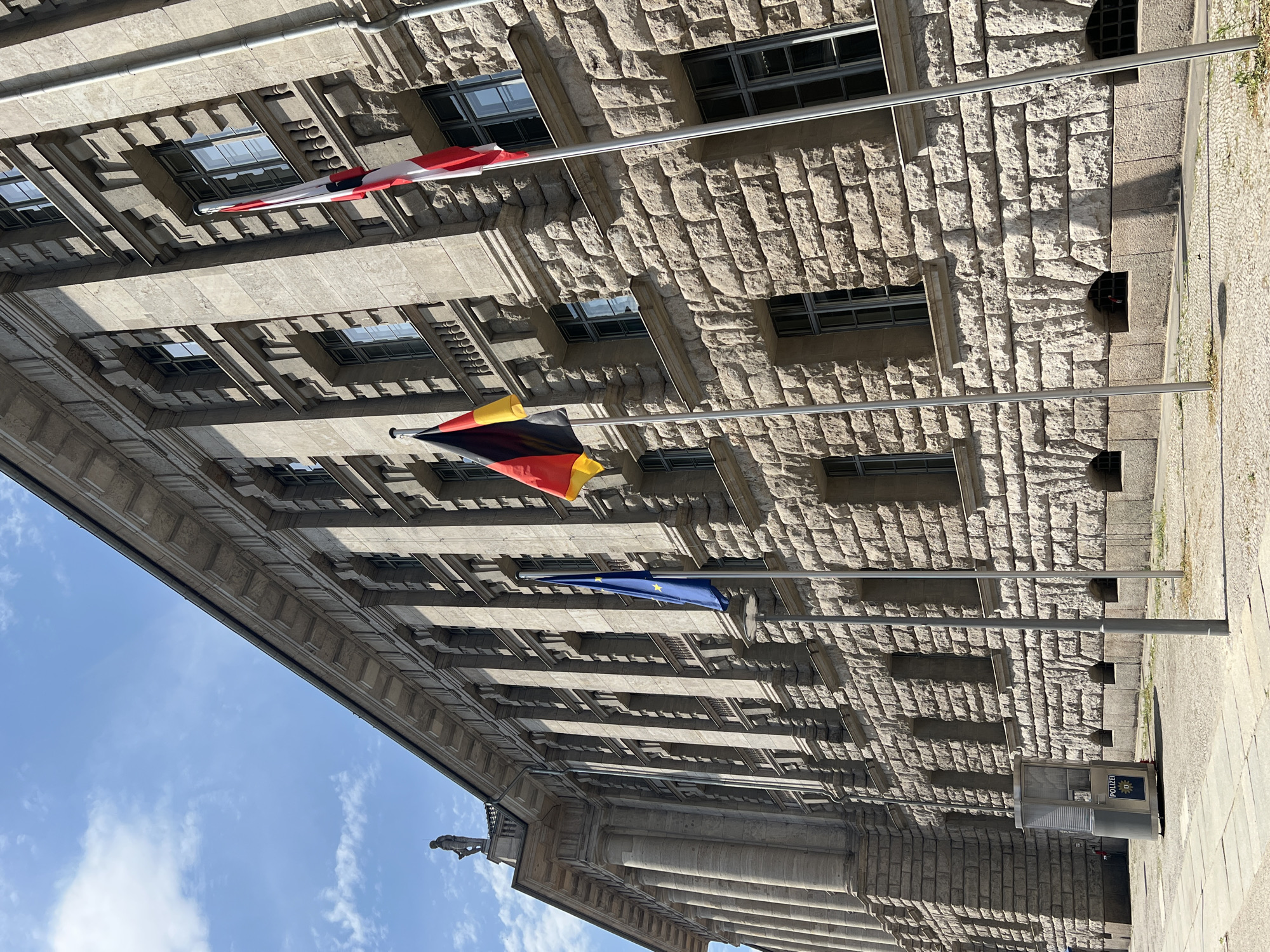 The image depicts the facade of a large, stone building with multiple rows of windows. Three flags are visible, suspended from poles attached to the building. The top flag is a red and white striped flag, likely a warning or signal flag. Below that is the German flag, featuring horizontal bands of black, red, and gold. The bottom flag is the blue European Union flag with its circle of yellow stars. The architecture of the building suggests a formal or governmental structure. The scene is set outdoors on a clear, sunny day, casting distinct shadows on the stone facade, indicating it is likely daytime. The building's rough-hewn stone blocks and the orderly arrangement of windows suggest an older, possibly historical, architectural style. The presence of the German and EU flags together implies the location is in Germany, possibly Berlin given the provided context. There is a small police booth visible in the lower right portion of the image with the word "POLIZEI" clearly legible, confirming the location is Germany and identifying the booth's function. The overall impression is one of officialdom and European unity.