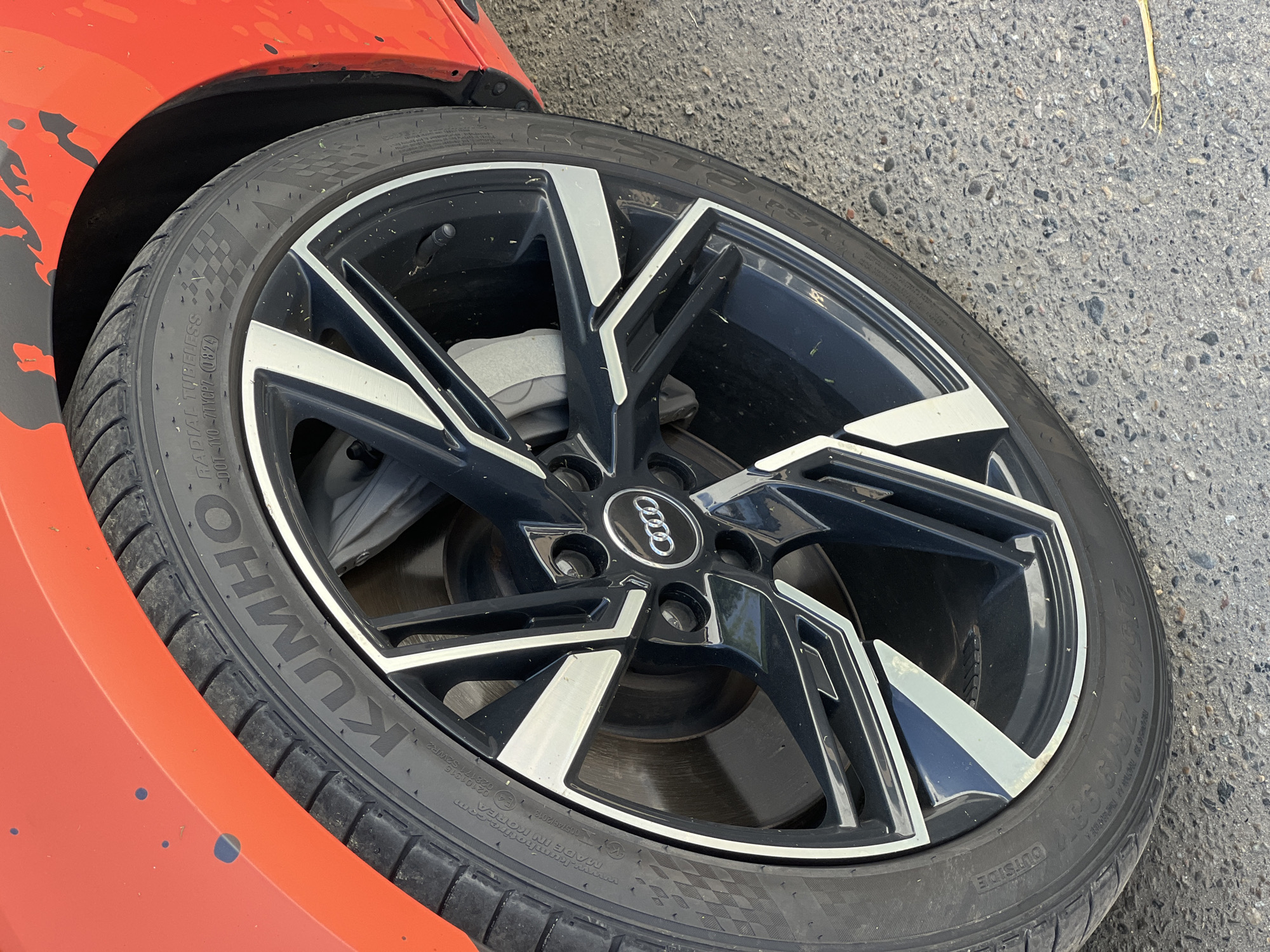 The image shows a close-up of a car's wheel and fender. The car is red, and the wheel is black with silver accents. The tire has "KUMHO" branding visible. The car appears to be parked on an asphalt surface. The image does not provide enough context to determine its specific location within Krasnodar, Russia. There are no people or actions depicted in the image.