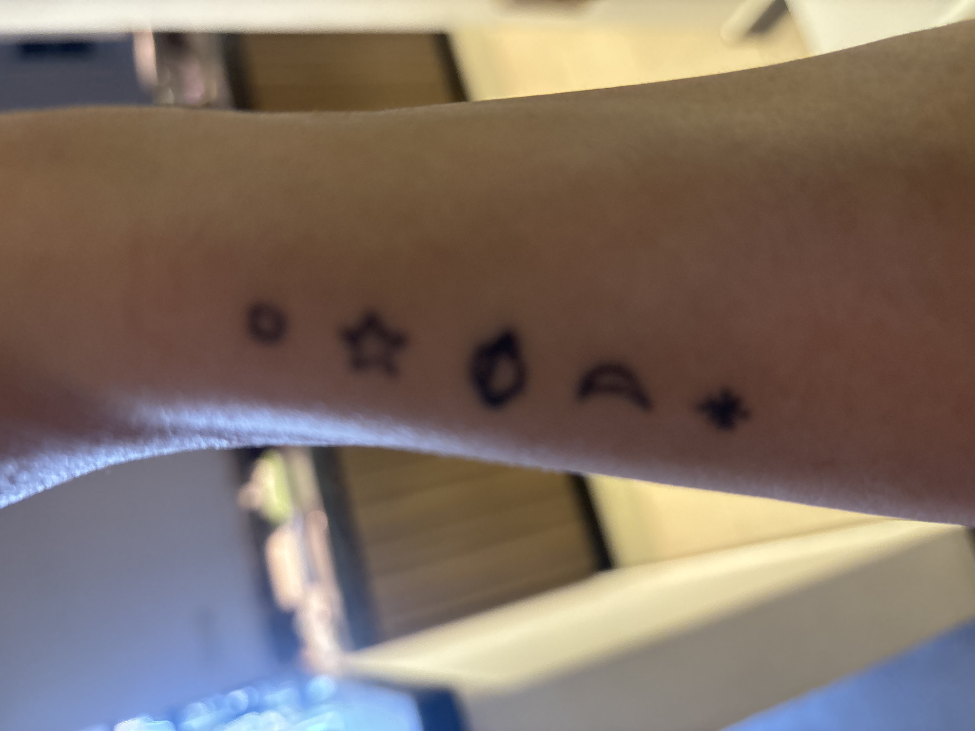 The image displays a close-up view of a person's forearm with several small, black stick-and-poke tattoos. From top to bottom, the tattoos are: a small circle, a five-pointed star, a shape resembling an eye or a leaf, a crescent moon, and a four-pointed star or asterisk. The skin appears to be light-toned. The background is blurred and shows what appears to be an indoor environment with wooden cabinets and a countertop, suggesting a kitchen or dining area. The lighting is somewhat dim, with a warm tone. No specific location in Dubai, United Arab Emirates can be determined from the visual information provided.