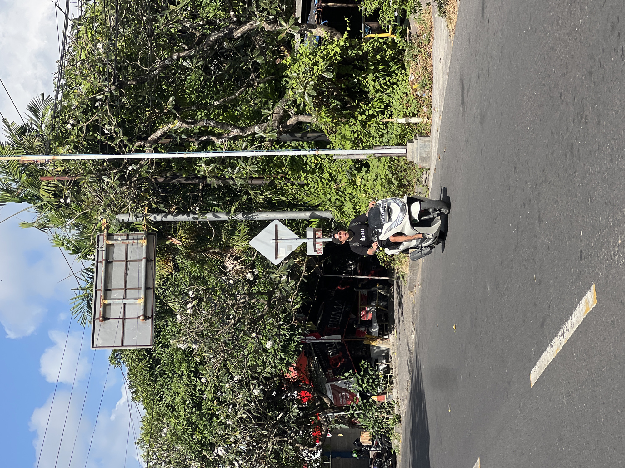 A man in a black t-shirt and black baseball cap rides a white scooter down a paved road in Kuta, Indonesia. The scooter has a license plate that reads "DKI 4378 ECR". The man appears to be looking forward. Behind him, lush green trees with white flowers are visible, with some palm fronds extending into the sky. A large, weathered billboard stands behind the trees, mounted on a metal frame. A traffic sign, shaped like a diamond, is also present. To the left of the road, there are parked scooters and a small, open-air shop with red and black awnings and signage. The sky is blue with scattered white clouds. The road has white dashed lines painted on it.