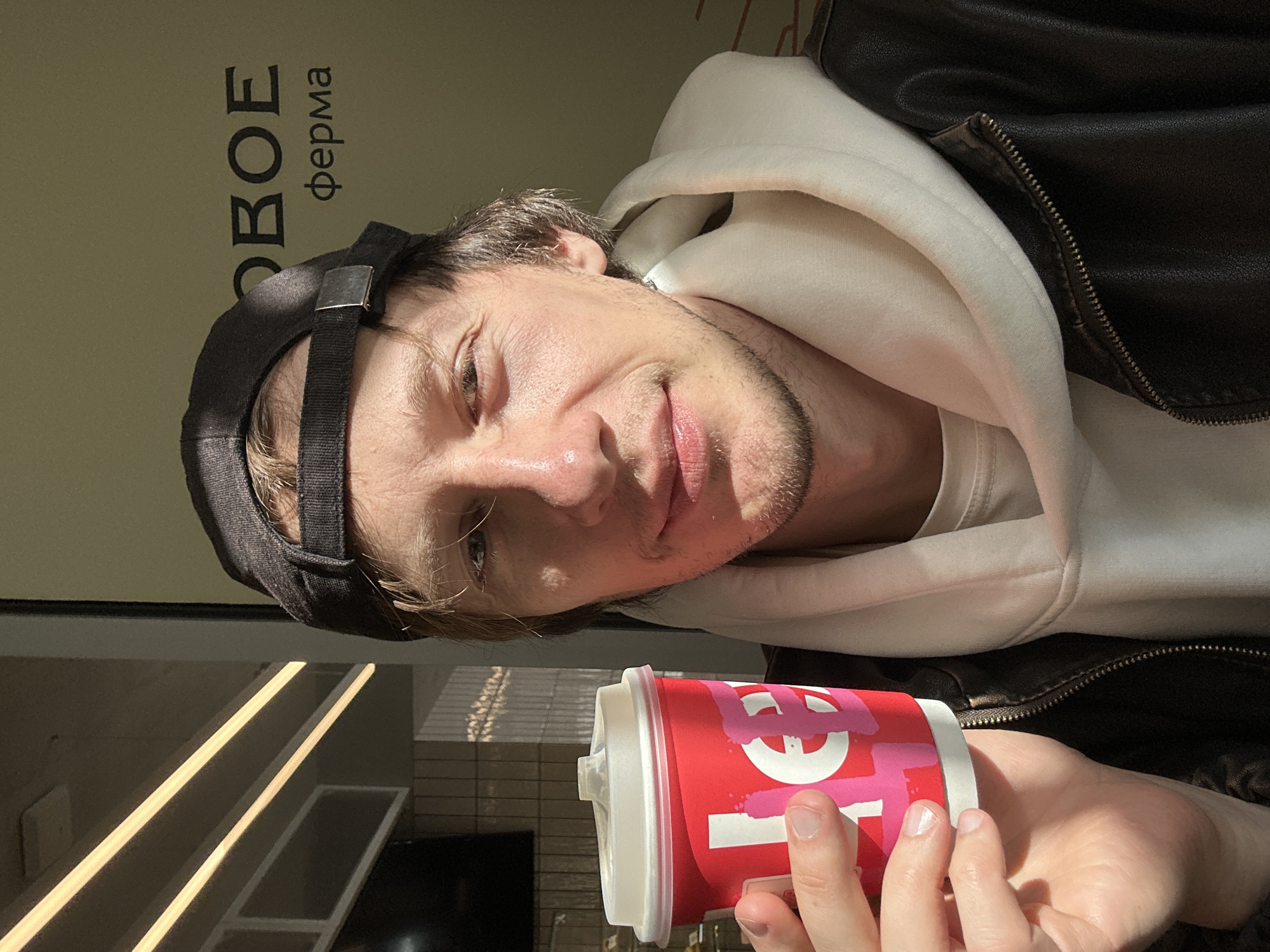 A fair-skinned man with light brown hair and stubble, wearing a black baseball cap, a white hoodie, and a dark brown leather jacket, is holding a red cup with a white lid and pink graffiti-style markings. The cup is held at the bottom in his hand. The man is looking at the camera with a slight smile. Behind him is a wall with Russian text that includes the word "ферма" (ferma, meaning "farm"). The background also shows light reflections, possibly from a window or reflective surface. Given the Cyrillic text, it is highly likely that this image was taken in Russia. Without additional evidence, identifying the exact location within Saint Petersburg is not possible.