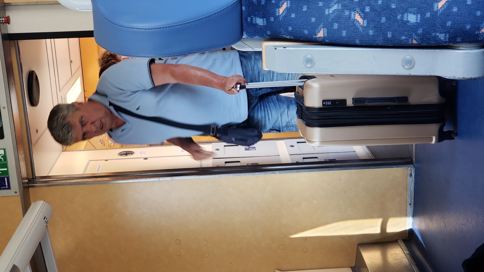 A middle-aged Caucasian man with graying hair and a light blue polo shirt and denim shorts stands on a train. He is carrying a dark blue shoulder bag and is holding the handle of a gold suitcase. The suitcase is positioned in front of him, with its wheels on the blue train floor. He is walking through a doorway with a metal frame. The train car interior features beige walls and blue seats visible in the foreground. Sunlight casts a shadow on the wall to the left. An emergency exit sign with green and white symbols is visible above the doorway.