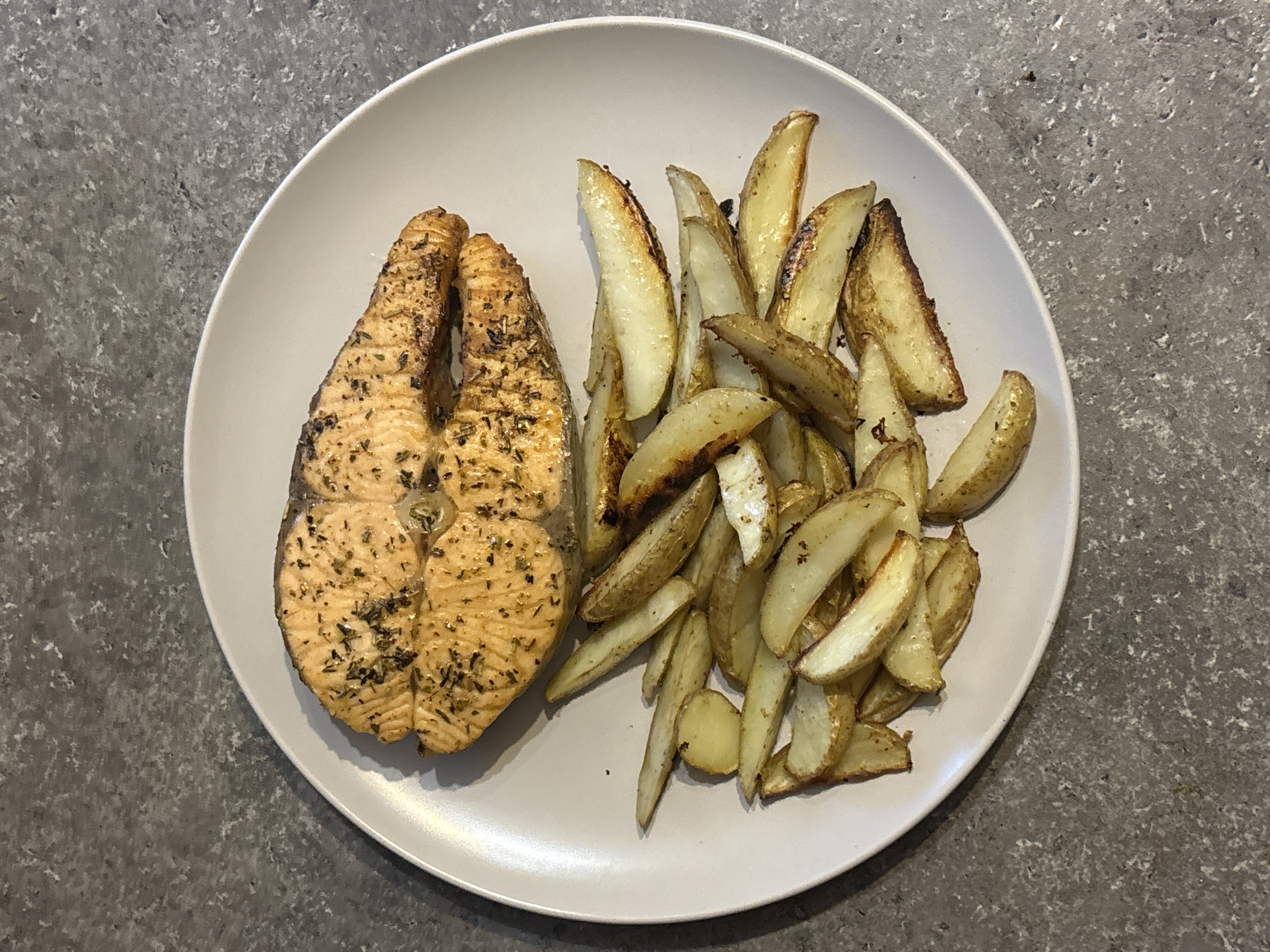 A top-down shot displays a light grey plate placed on a grey countertop, featuring a meal consisting of a grilled salmon steak and a heap of potato wedges. The salmon is situated on the left side of the plate, seasoned with herbs. Potato wedges are piled up on the right side, exhibiting a golden-brown color. The plate's surface is mostly clean except for the food items. The countertop has a speckled texture. The image does not contain any people, actions, or interactions. It is impossible to tell whether this image was taken in the city of Верхнеказымский, Russia.